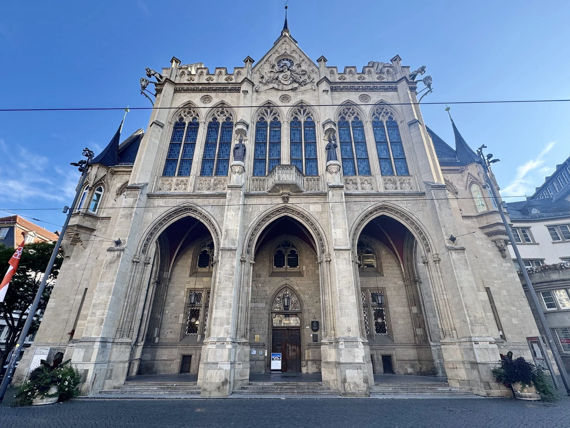Front view of a Gothic-style church with tall pointed arches, large stained glass windows, decorative statues, and a central clock, with a clear blue sky in the background.