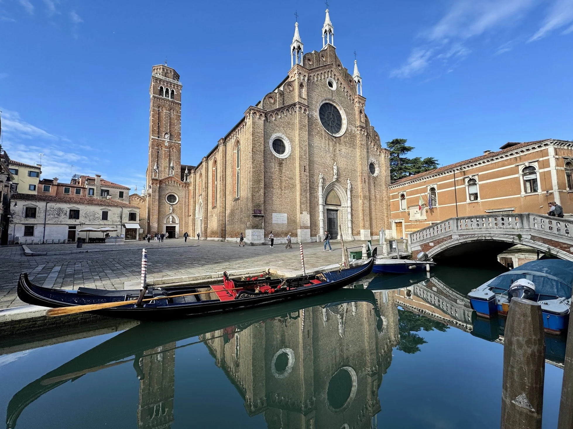 A historic church with a tall bell tower, reflected in the calm water of a canal with gondolas, set in a sunny day with clear blue sky and a few clouds.