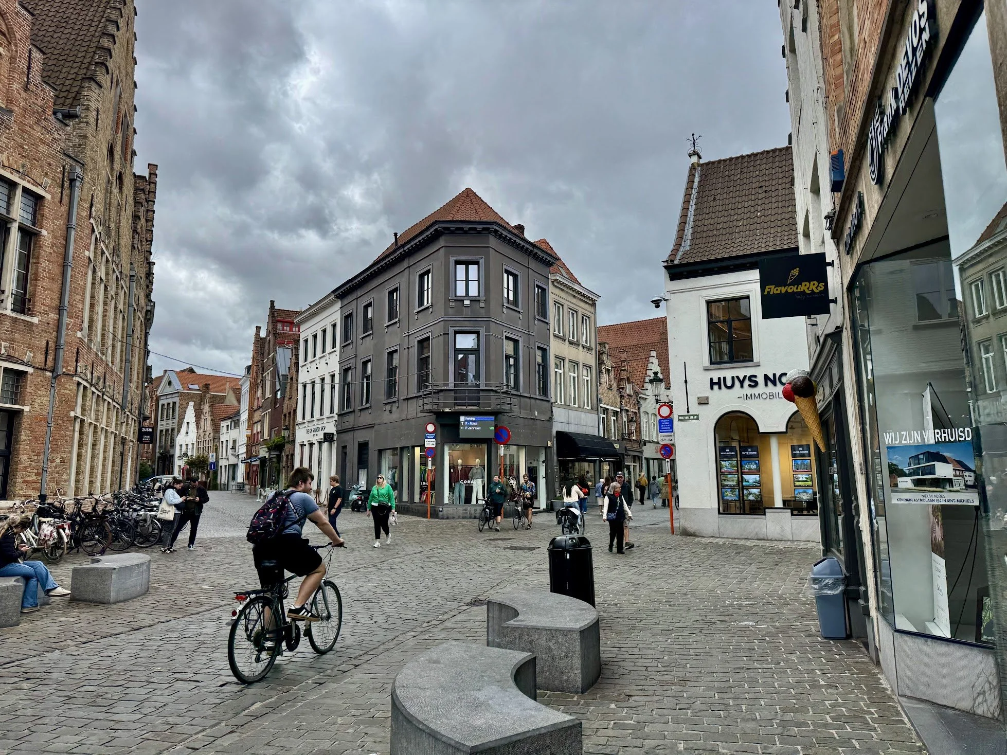 A busy urban street scene with several pedestrians, bicycles, and storefronts under cloudy skies. Buildings feature brick and modern facades.