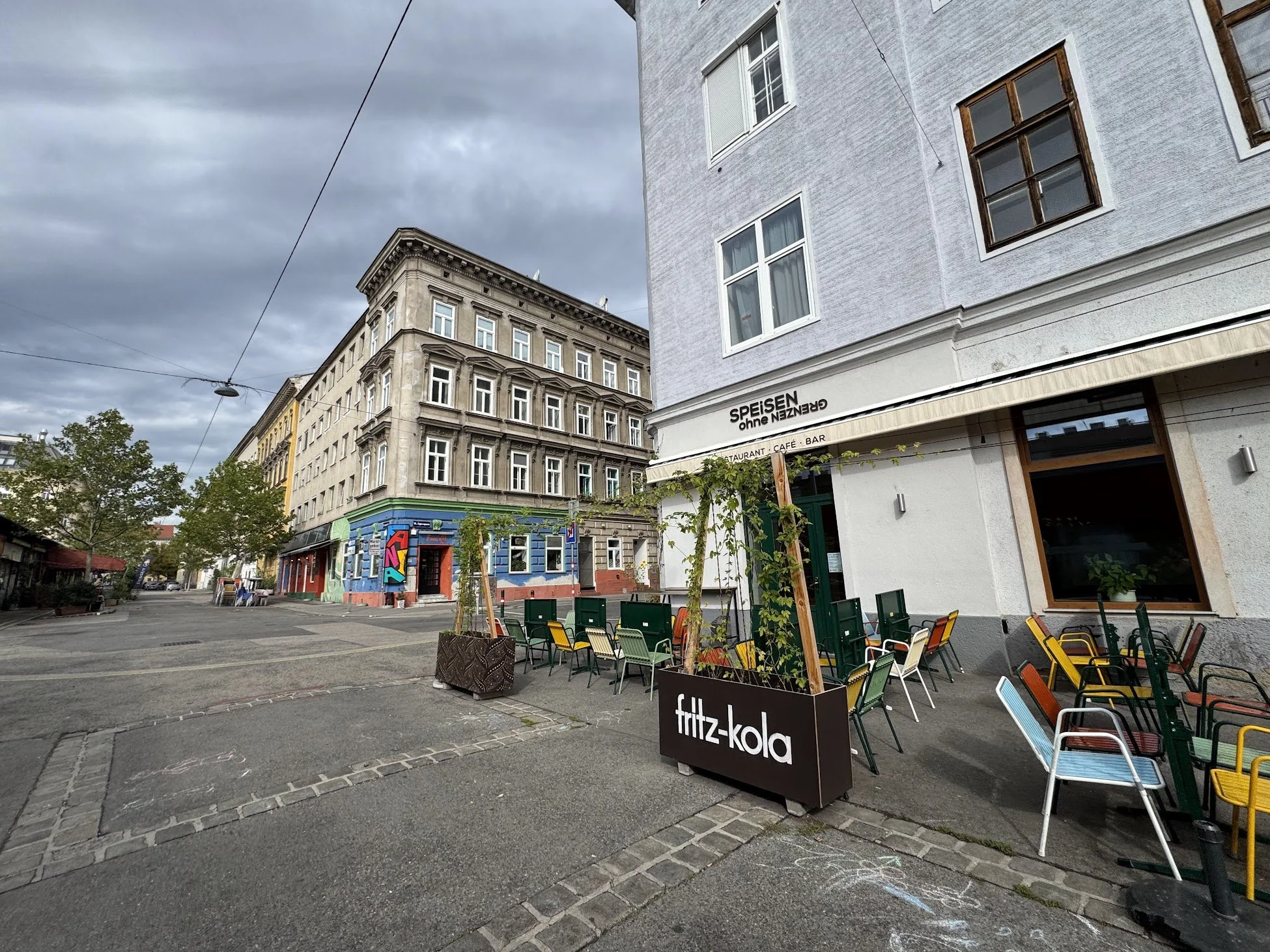 City street scene with outdoor seating, colorful chairs, a sign for Fritz-Kola, and adjacent buildings under cloudy sky.