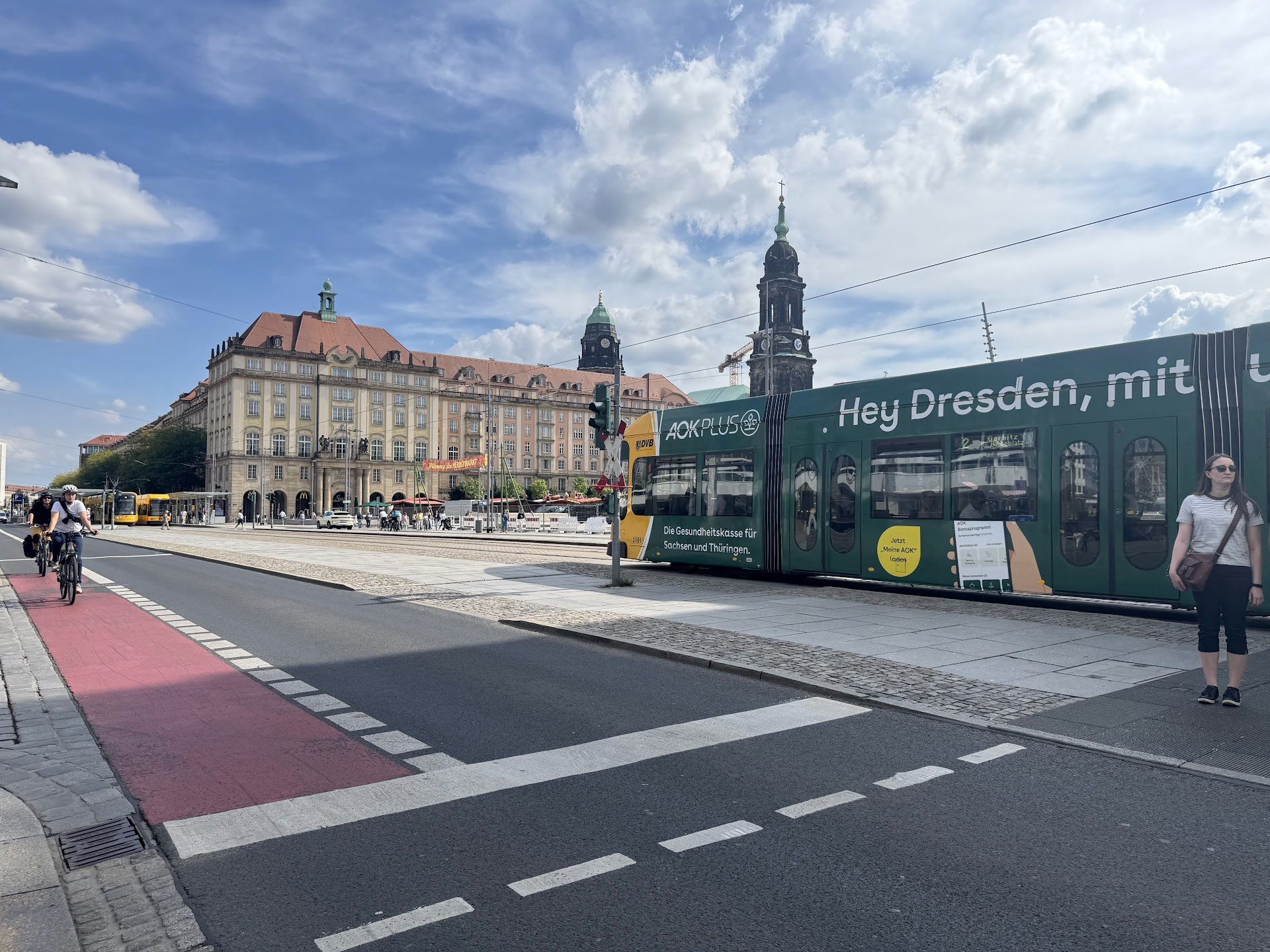 A city street scene with a historic building with towers and a clock in the background, a green tram with advertising, cyclists on a red bike lane, and pedestrians crossing the street under a partly cloudy sky.