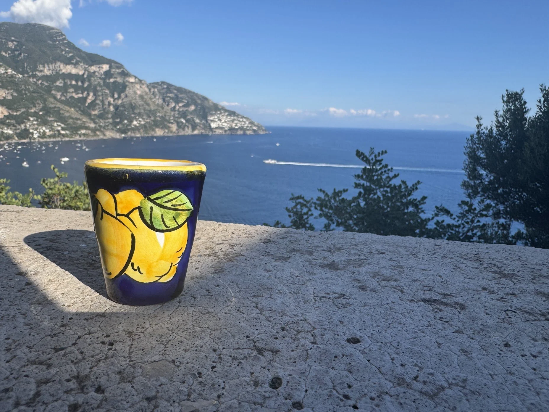 A colorful ceramic mug featuring a lemon design, placed on a stone surface overlooking a scenic coastal landscape with blue water, boats, green trees, and mountains in the background.