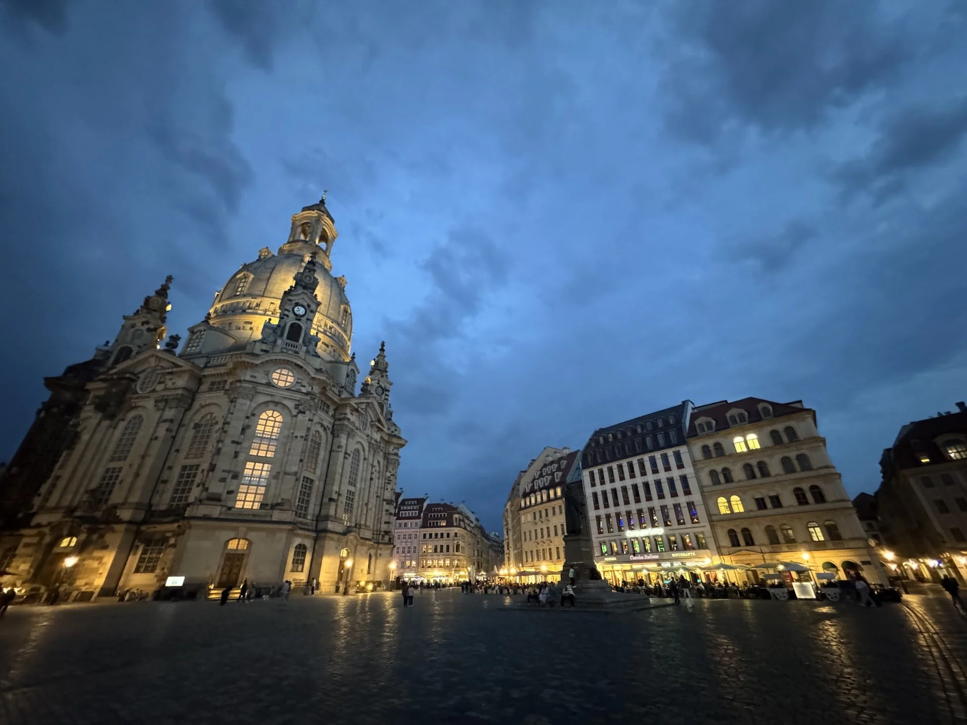 A historic European city square during twilight with a large baroque church and surrounding buildings illuminated by streetlights, crowded with people.