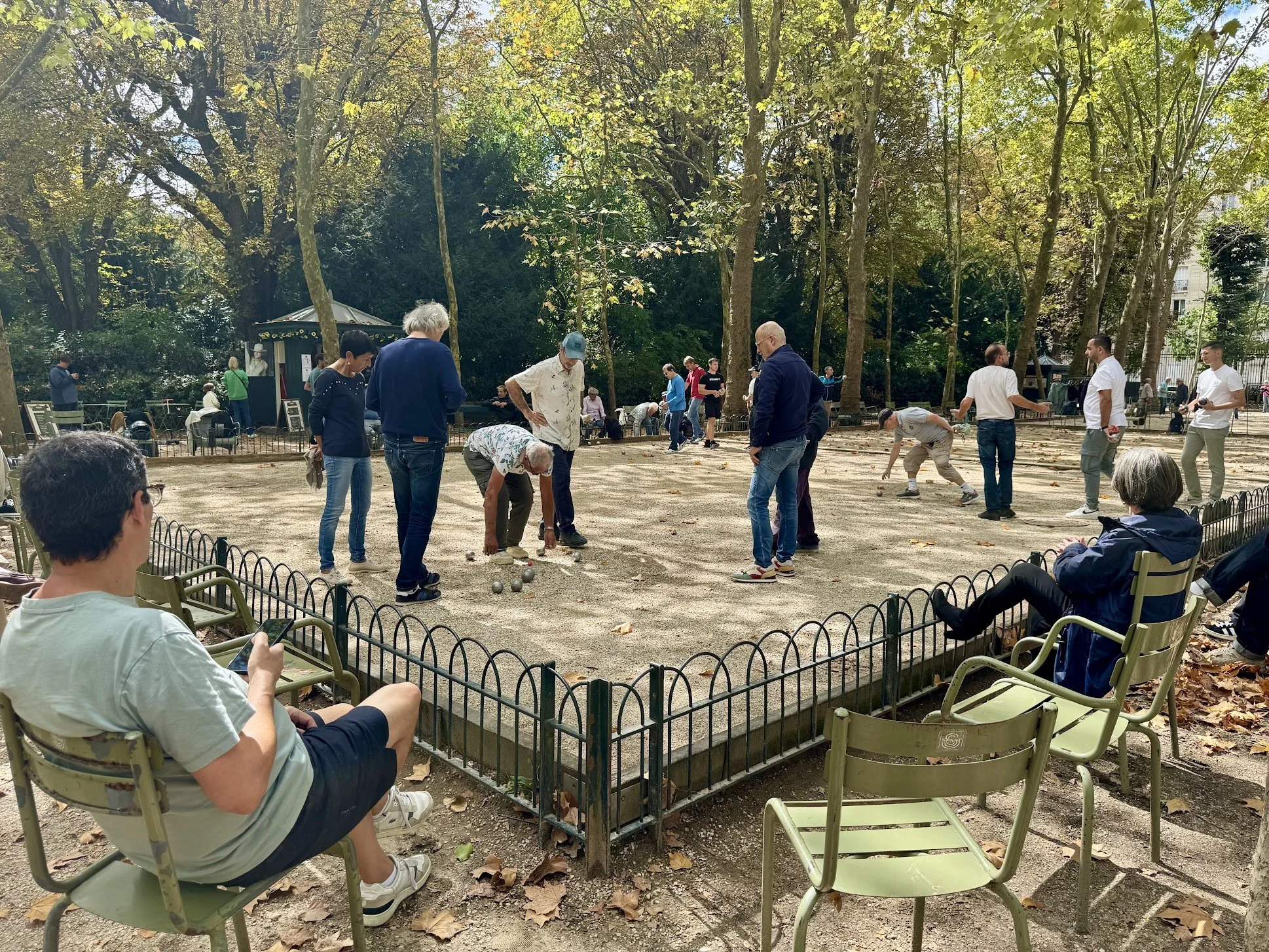 People playing pétanque on a dirt court in a park surrounded by trees with green and some yellow leaves, with a few spectators sitting on green chairs.