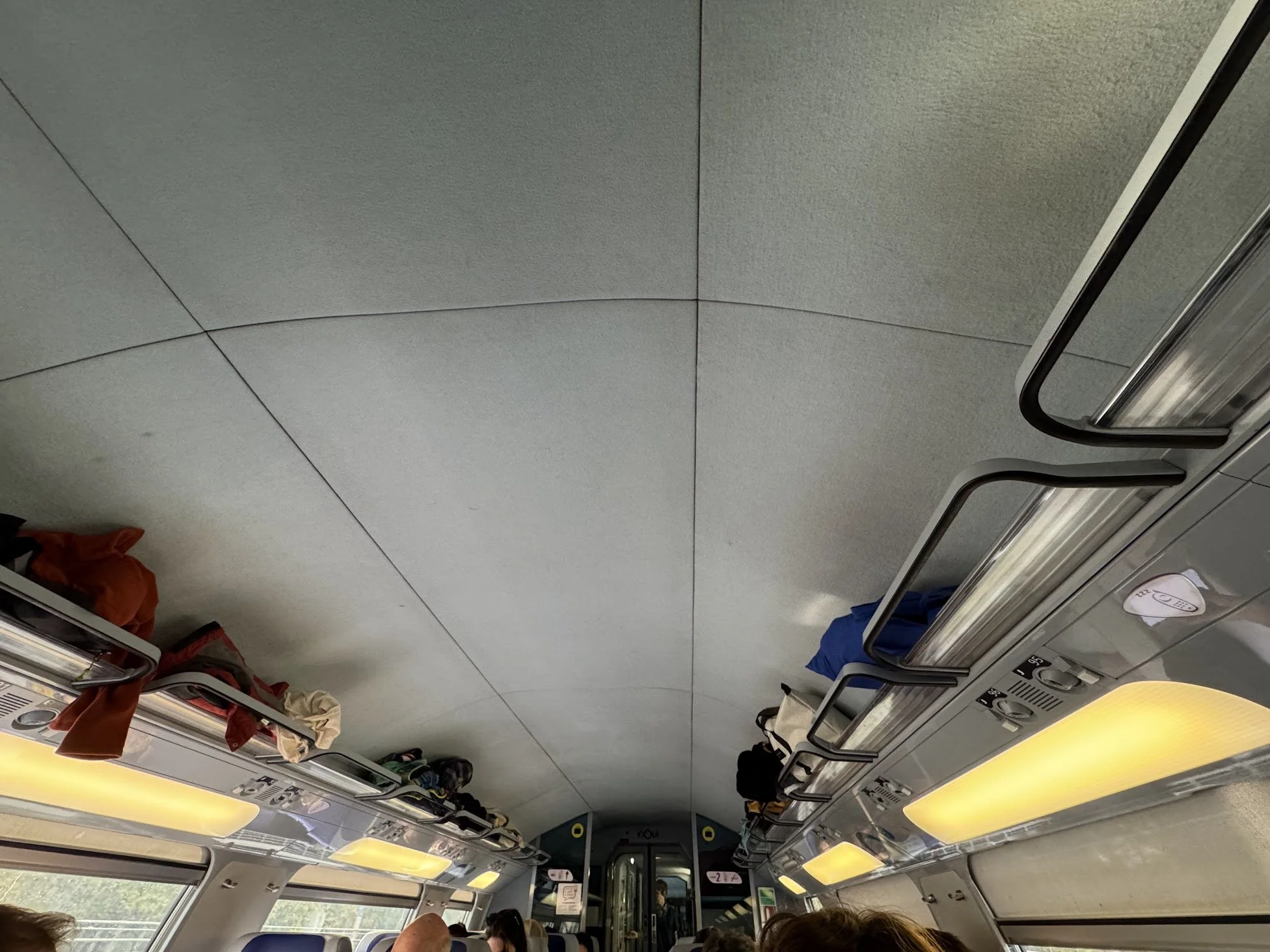 Interior of a passenger train, showing overhead storage compartments with clothes and bags, yellow-lit ceiling, windows with passengers seated below, and partially open doors at the end of the car.
