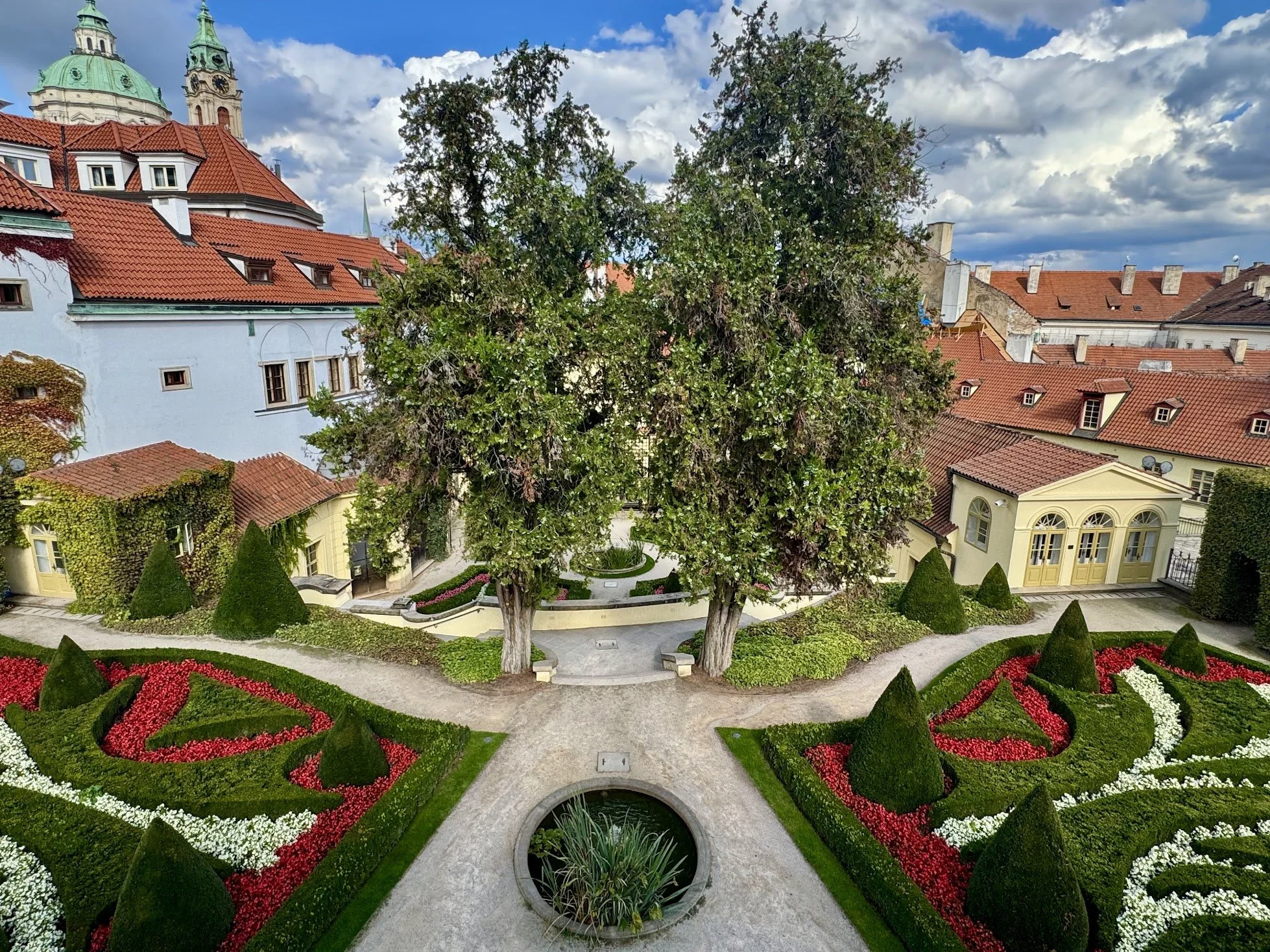 A courtyard garden with geometric flower beds filled with red, white, and pink flowers, trimmed bushes, and a decorative water fountain, surrounded by historic buildings with red-tiled roofs and a large tree.