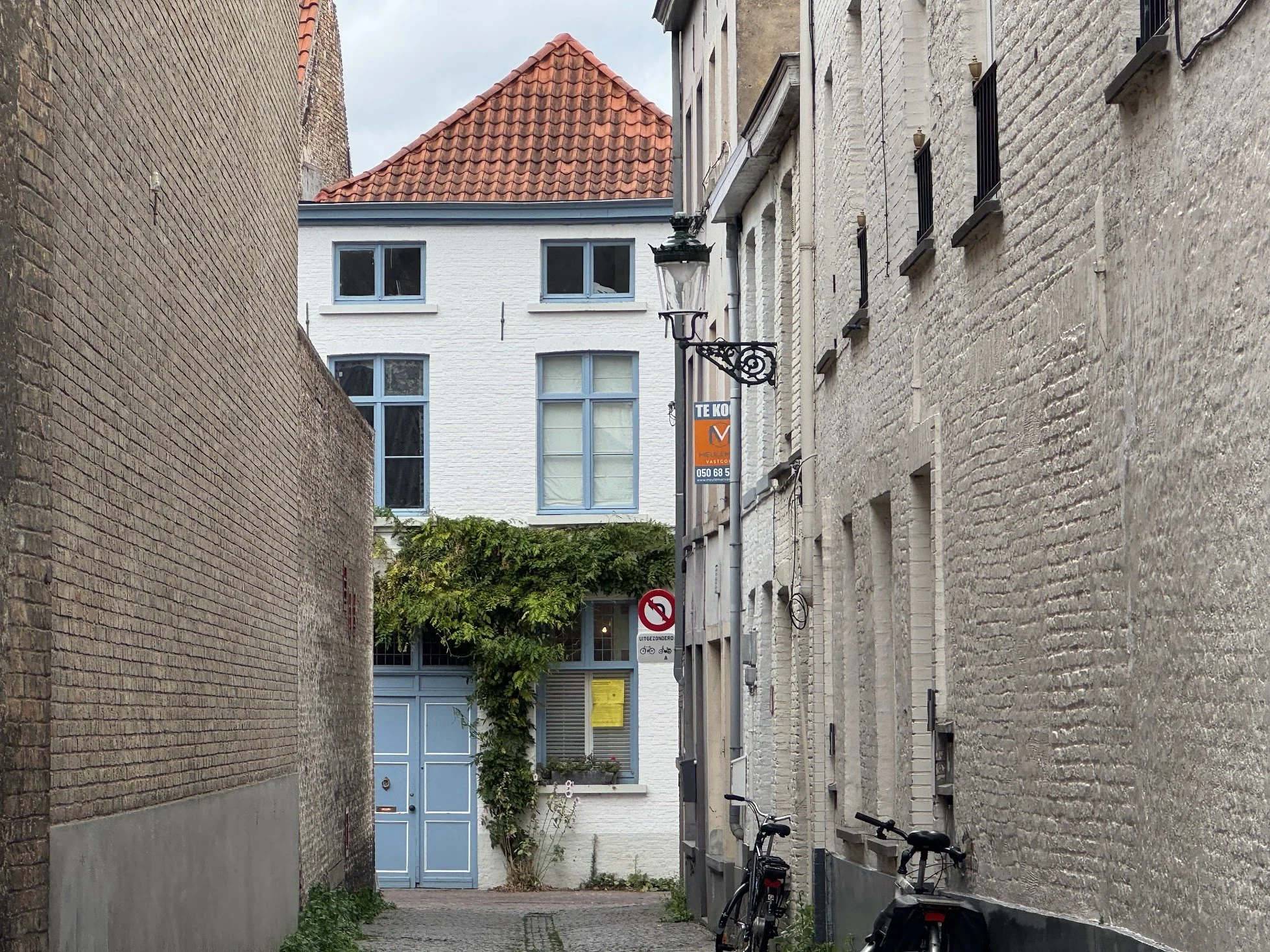 A narrow alleyway with brick walls on either side, industrial-style lamp posts, bicycles parked on the right, a green leafy tree at the end, and a building with white walls and blue-framed windows in the background.