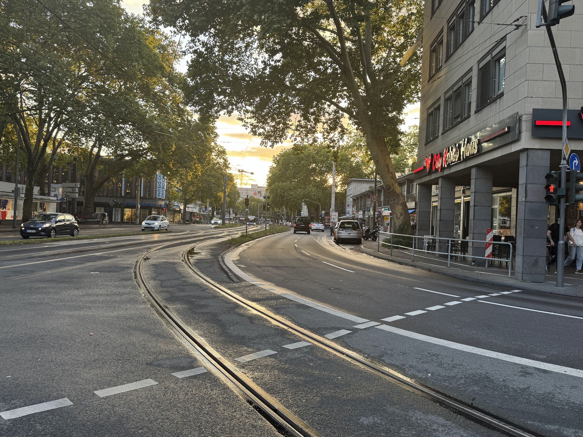 Street scene at sunset with tram tracks, cars, pedestrians, and a large building with illuminated signage in an urban area.