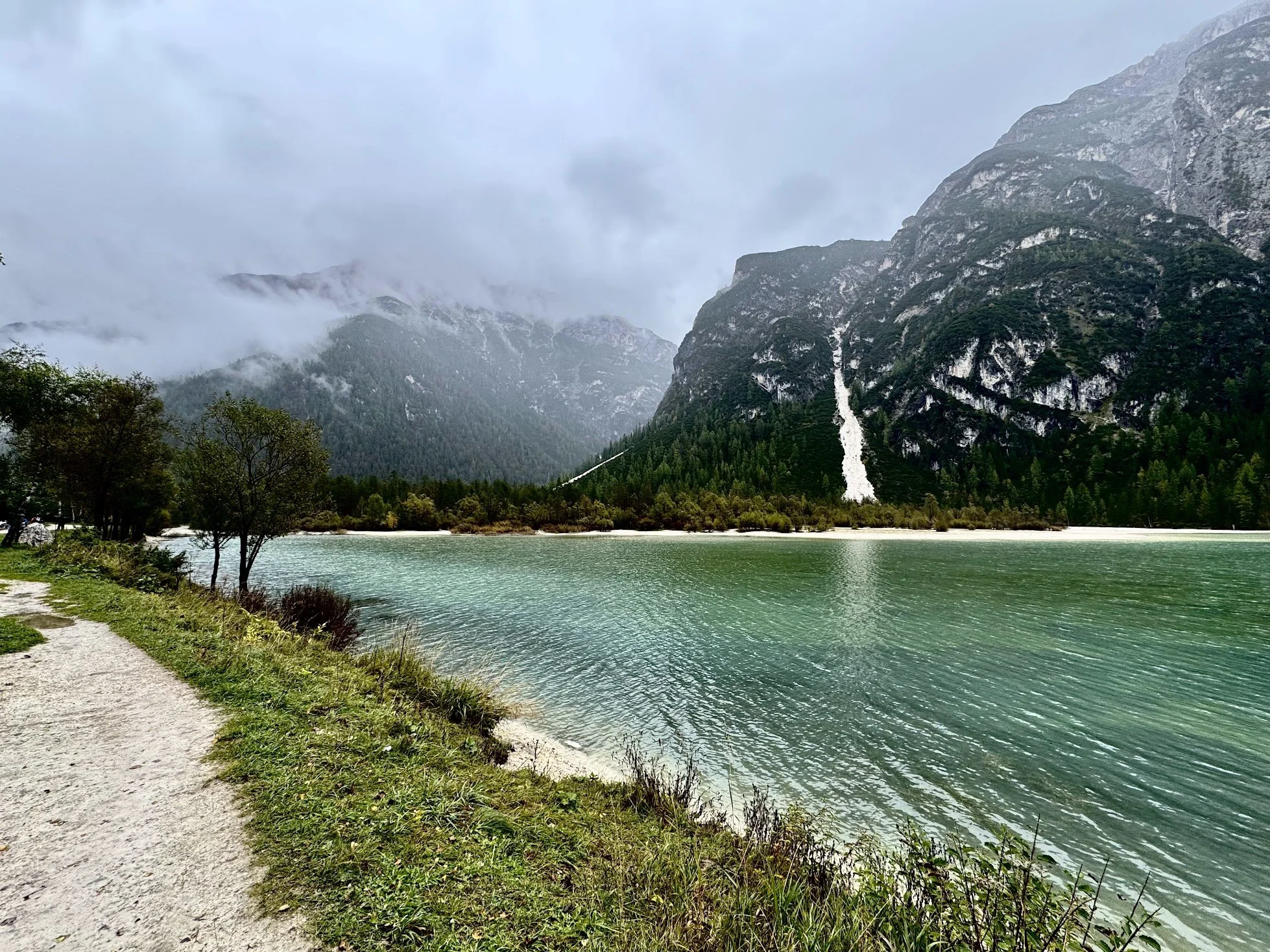 A lake with greenish water, surrounded by lush greenery and trees. In the background, large rocky mountains partially covered in fog or low clouds, with the sky overcast.