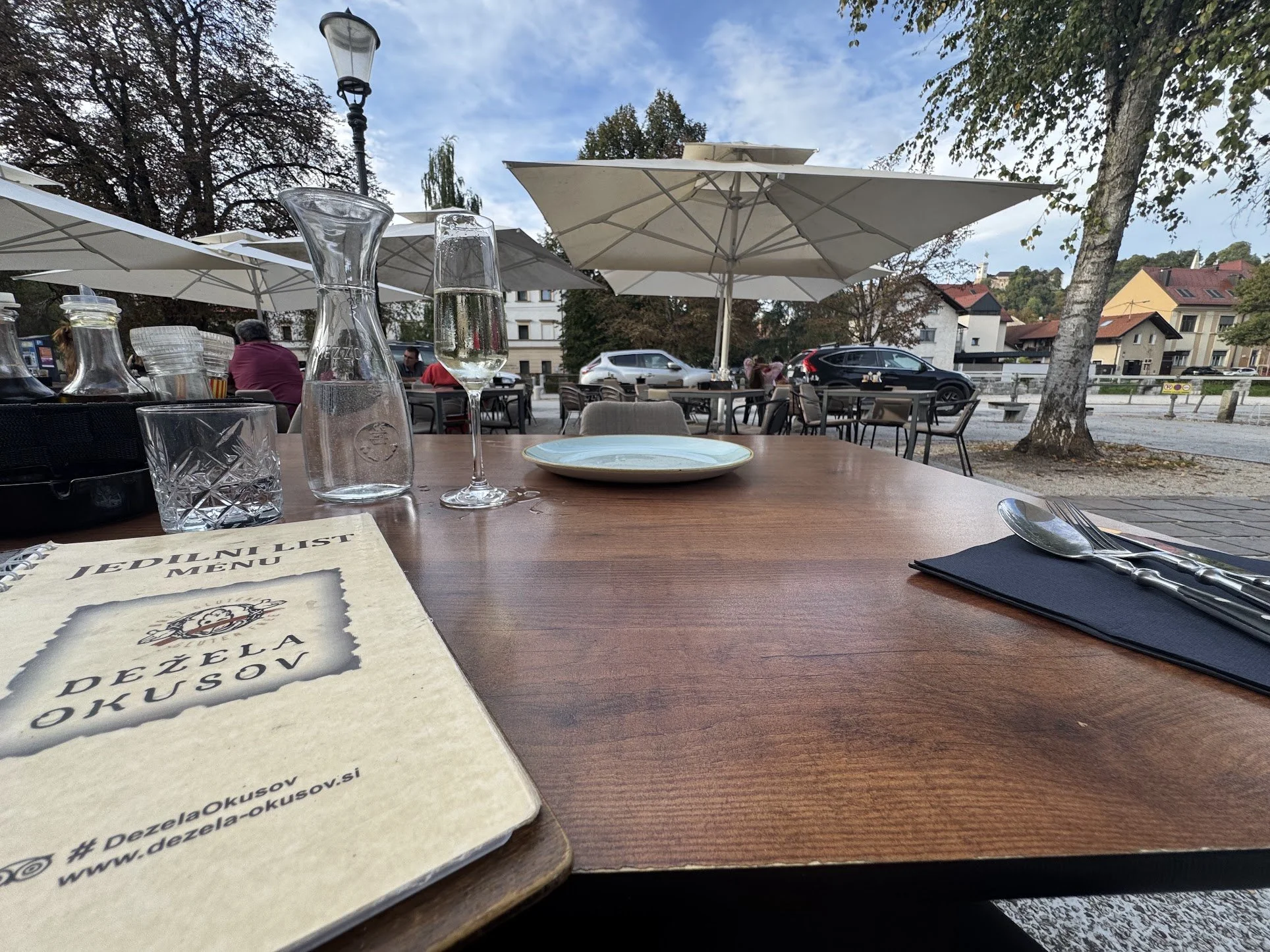 Outdoor restaurant patio with tables, umbrellas, and a view of parked cars and residential buildings. On the table in the foreground, there are glasses, a notepad, and plates.