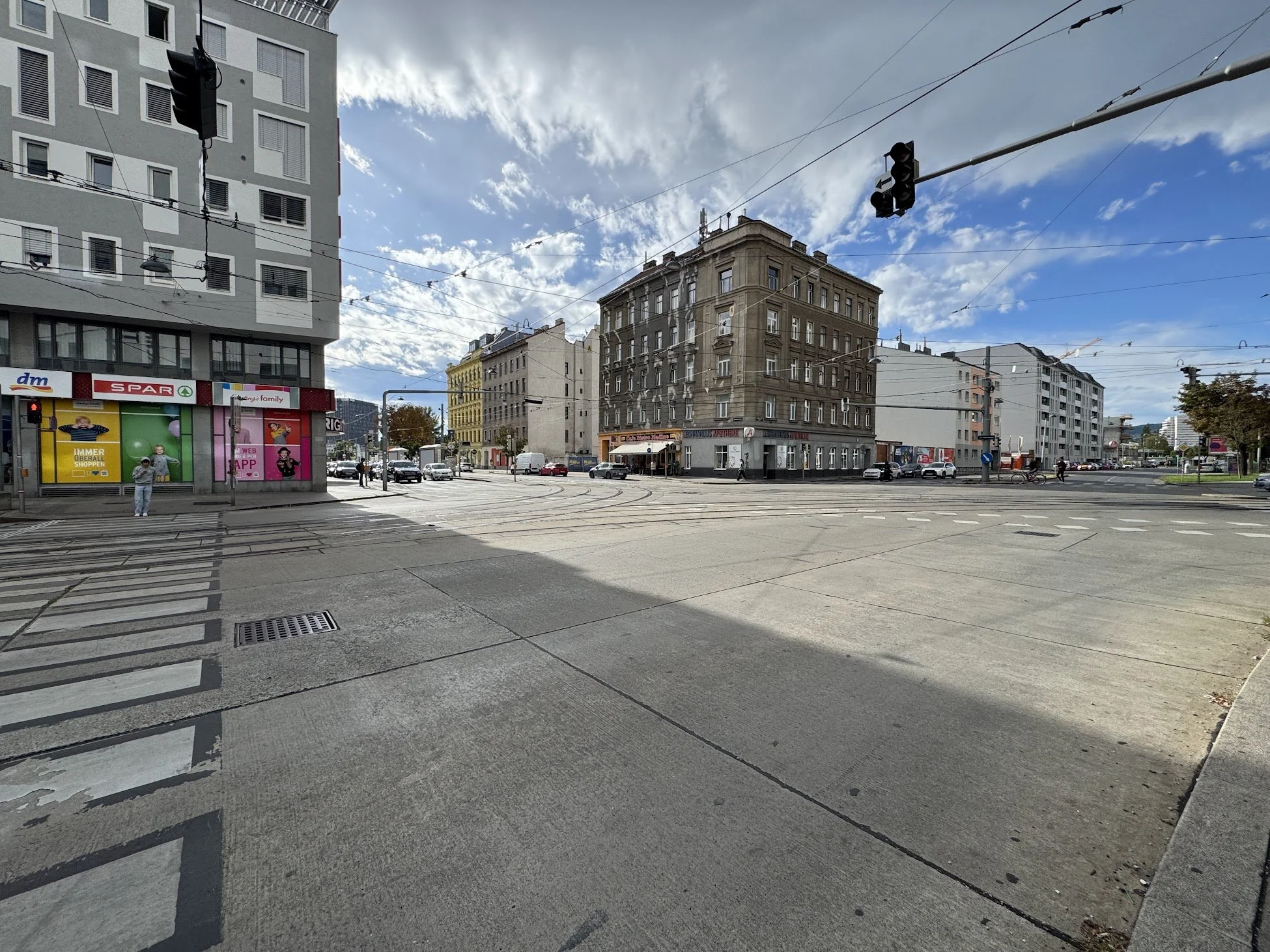 A city street scene with a crosswalk, tram tracks, traffic lights, and tall buildings. People are walking or waiting at the crosswalk on a partly cloudy day.