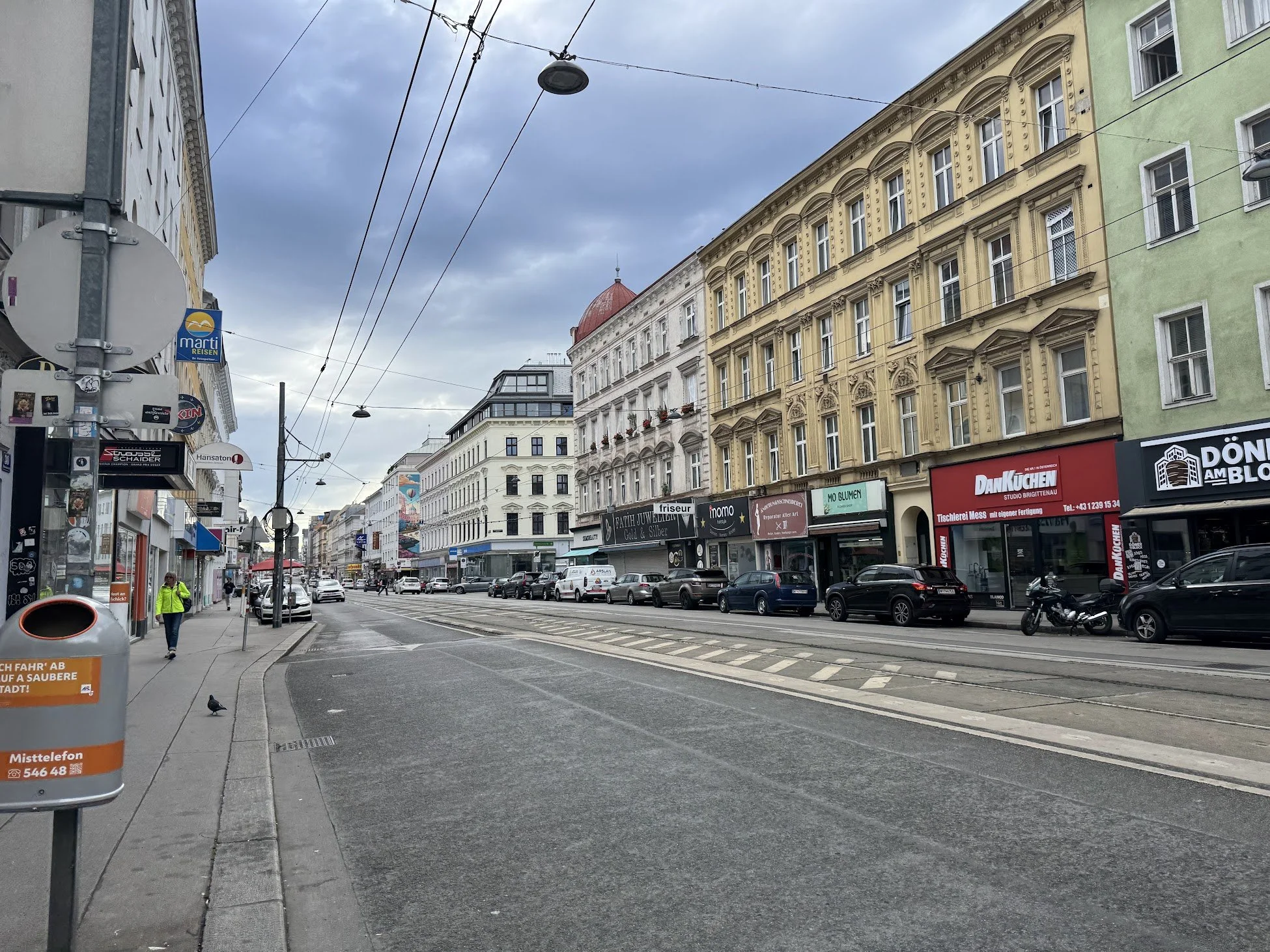 A city street with historic buildings, parked cars, and tram lines overhead under cloudy skies.