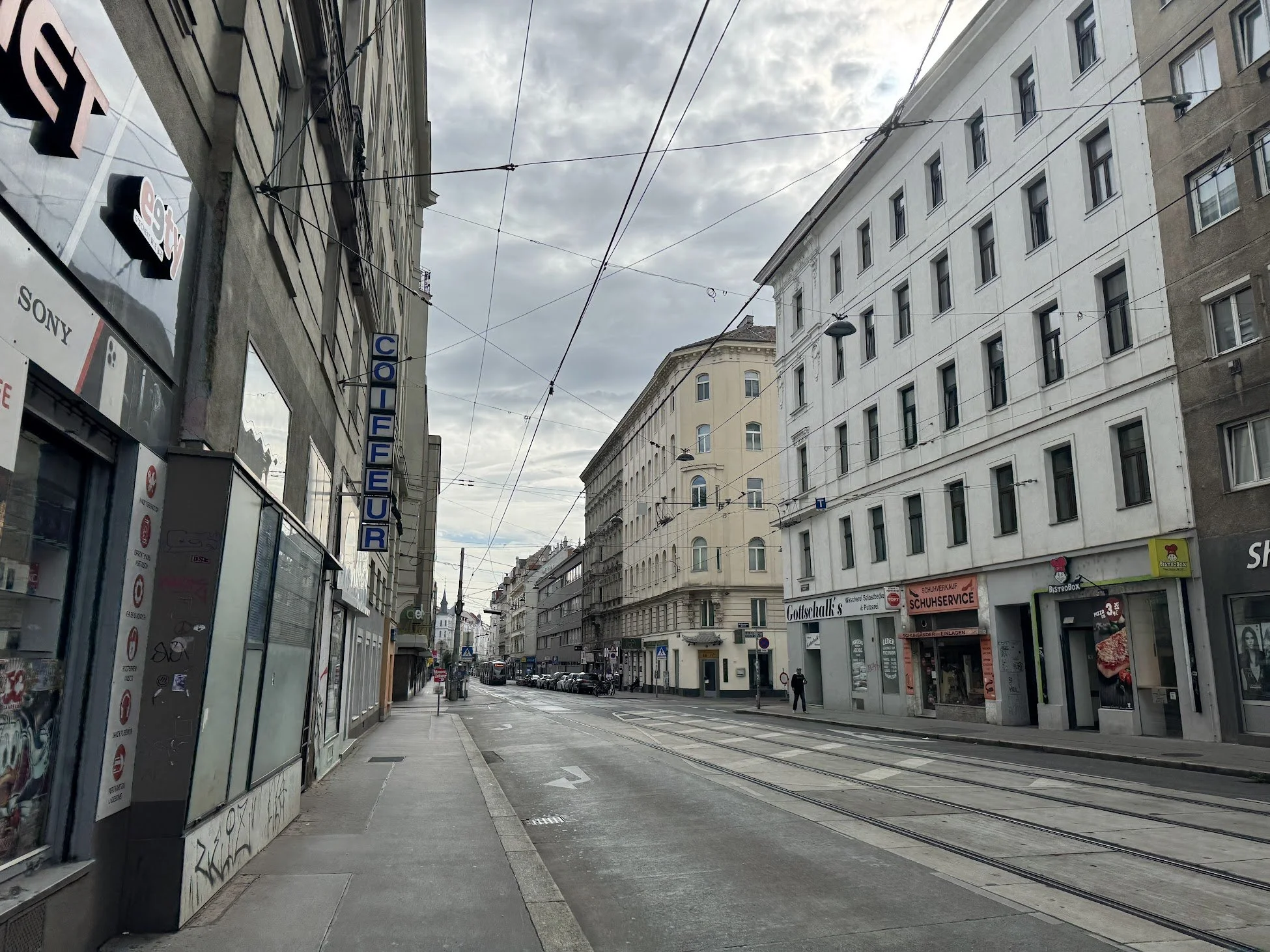 A city street with tram tracks and overhead wires, surrounded by tall buildings with a cloudy sky overhead.