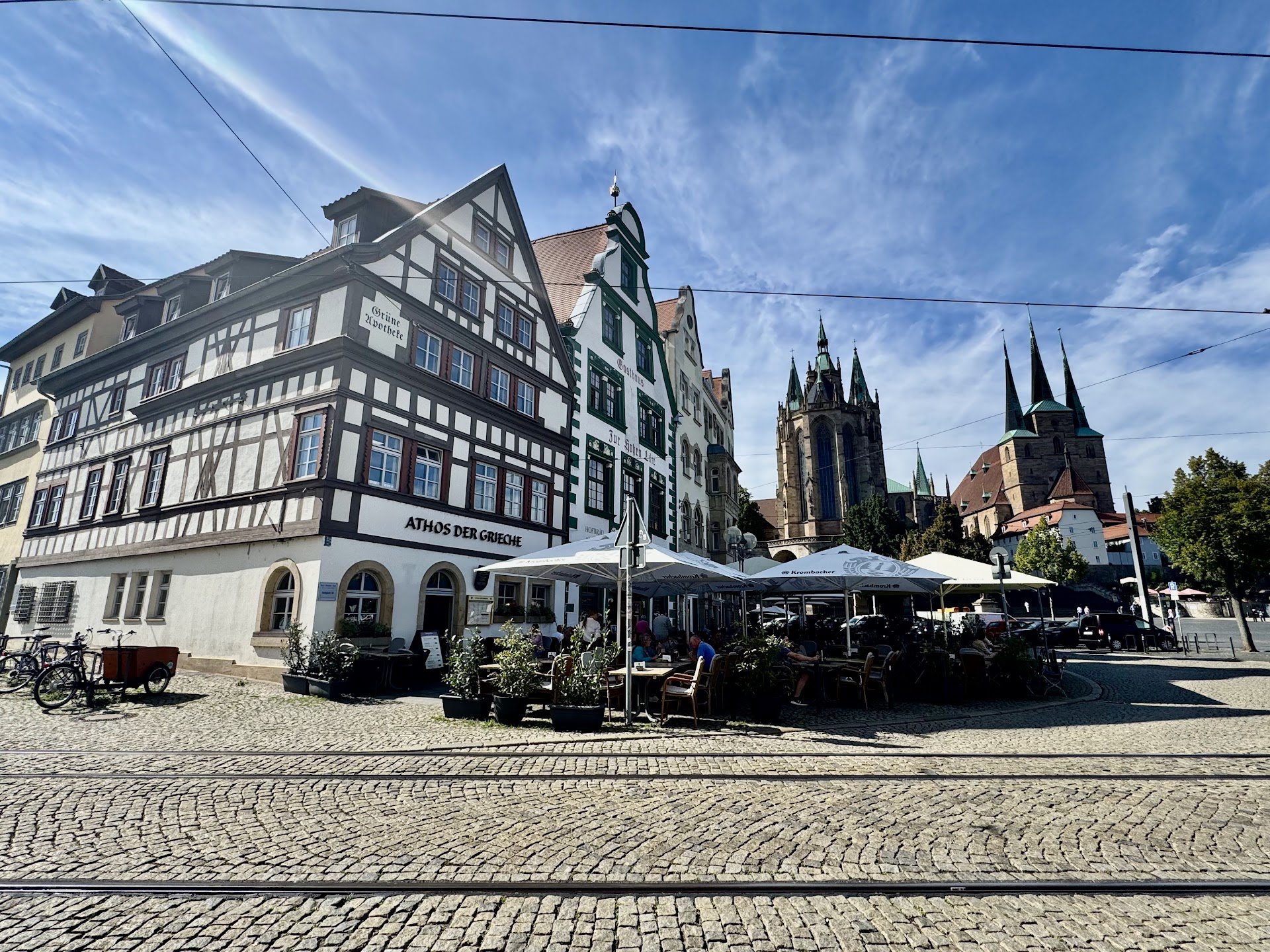 A European city square with historic buildings, outdoor cafe seating, and a church with tall spires in the background under a blue sky.