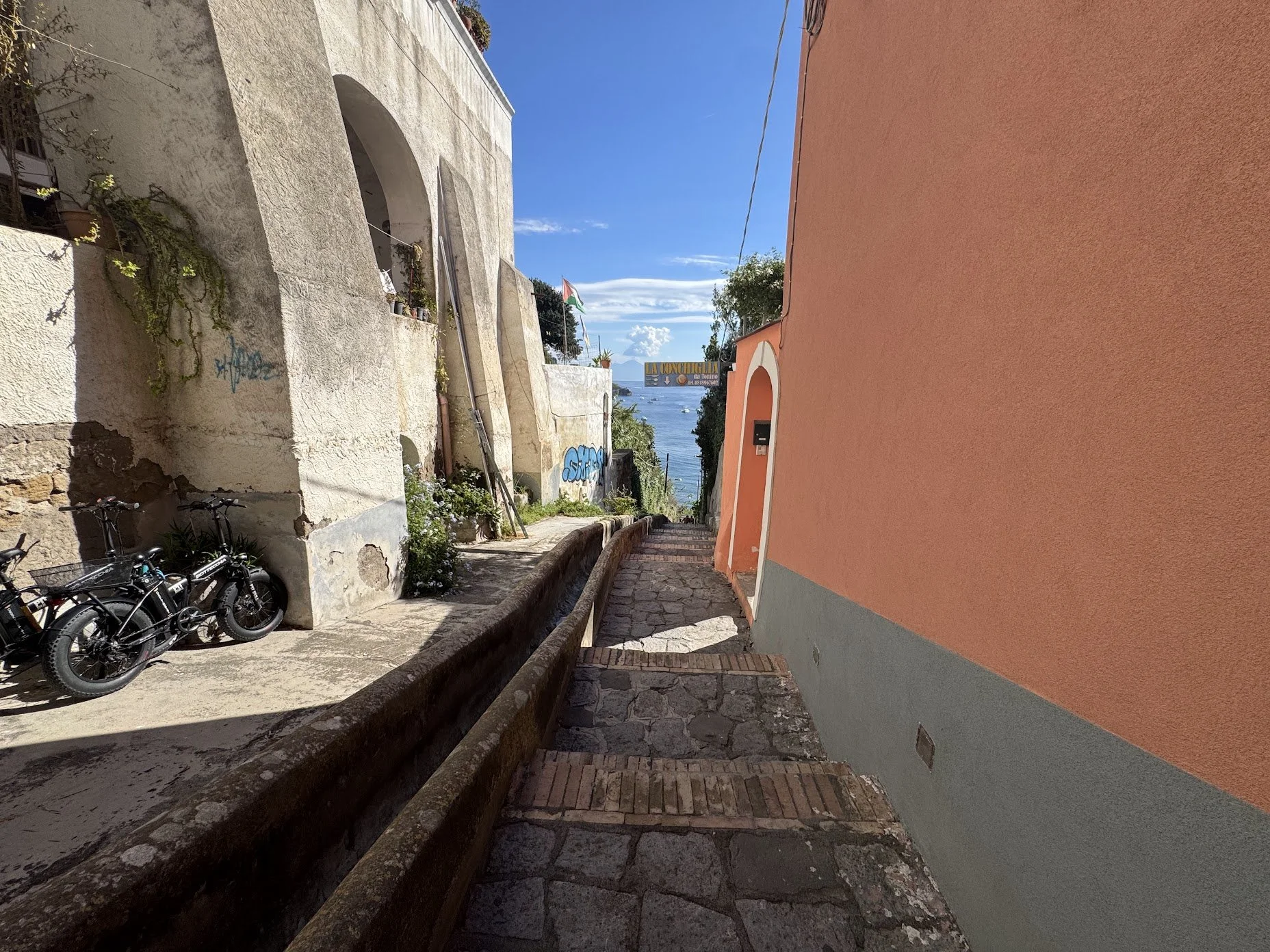 A narrow cobblestone street on a hillside overlooking water, with two buildings on either side, one painted orange and the other with worn white walls and graffiti. Several bicycles are parked next to the white building. The sky is clear with a few c