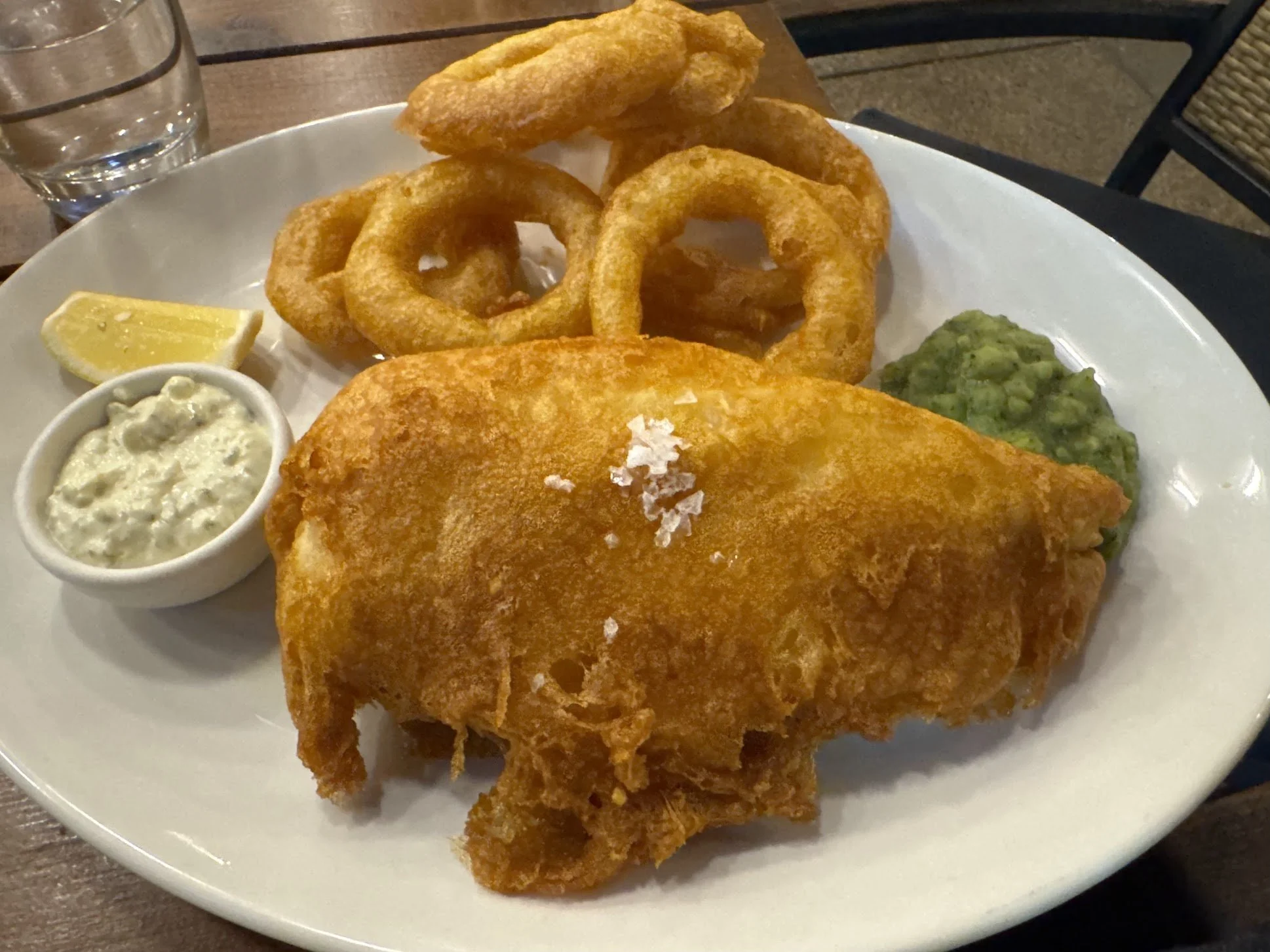 Plate of battered deep-fried fish fillet, onion rings, and a side of tartar sauce and a lemon wedge, with a glass of water in the background.
