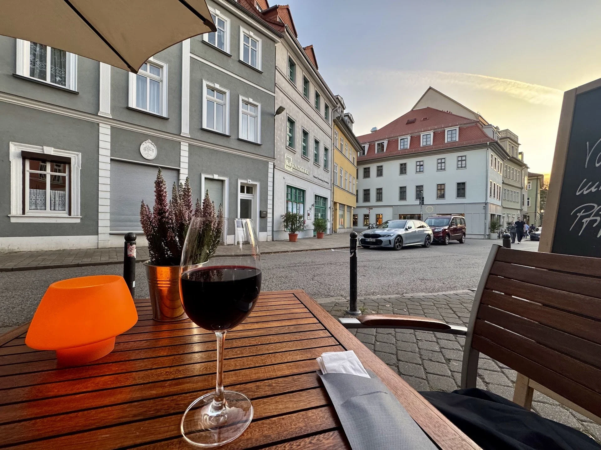 A table at an outdoor cafe with a glass of red wine, an orange hat, and a napkin on a wooden table. In the background, a street scene with parked cars and multi-story buildings under a clear sky with the sun setting.