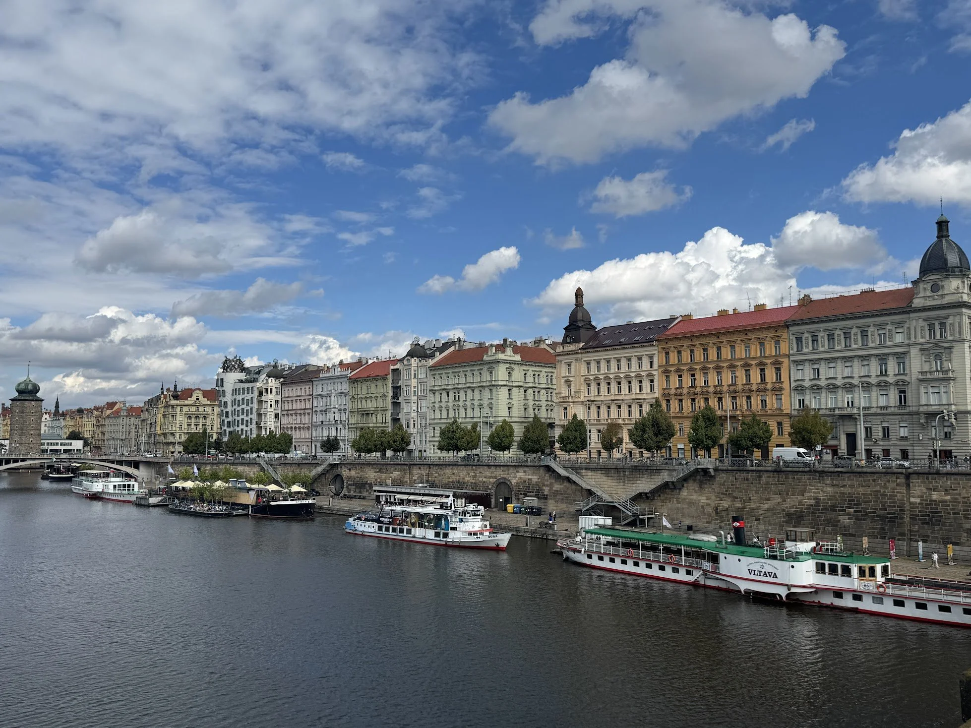 A river with boats docked along a historic European city with colorful buildings and church towers under a blue sky with fluffy clouds.