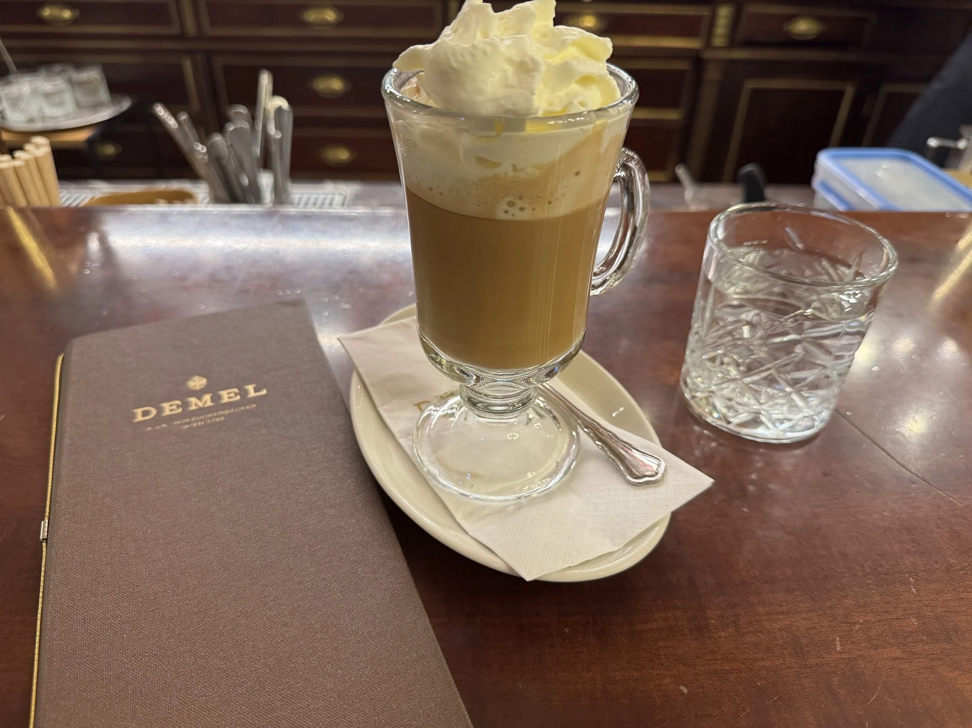 A glass mug filled with coffee topped with whipped cream, placed on a saucer with a teaspoon. Next to the mug is a glass of water, and a closed menu titled "DANEL DEMEL" rests on the table. The background shows a wooden counter with drawers and vario