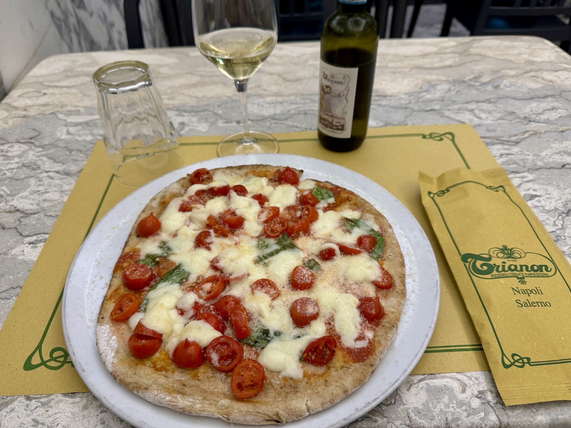 A pizza with cherry tomatoes, basil, and cheese on a white plate on a yellow and green tray, with a glass of white wine and a bottle of wine on a marble table.
