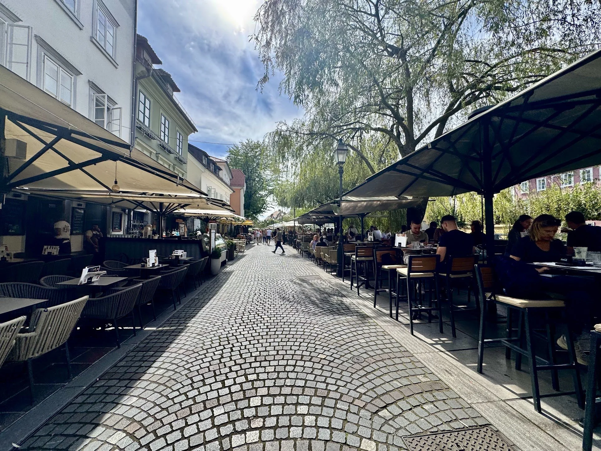 Outdoor dining area with tables and chairs under large umbrellas on a cobblestone street, with trees and buildings in the background.