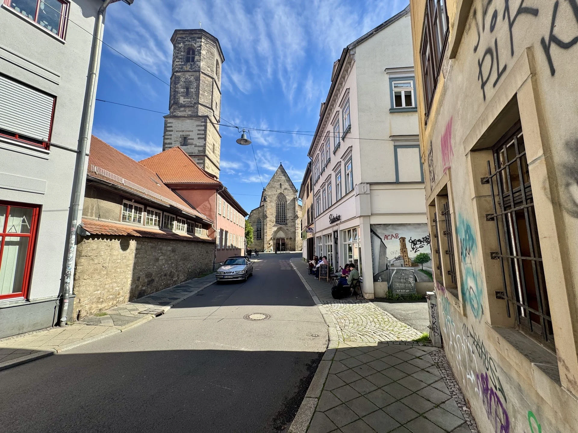 A street view with historic church tower under blue sky, surrounded by colorful buildings and people sitting outside