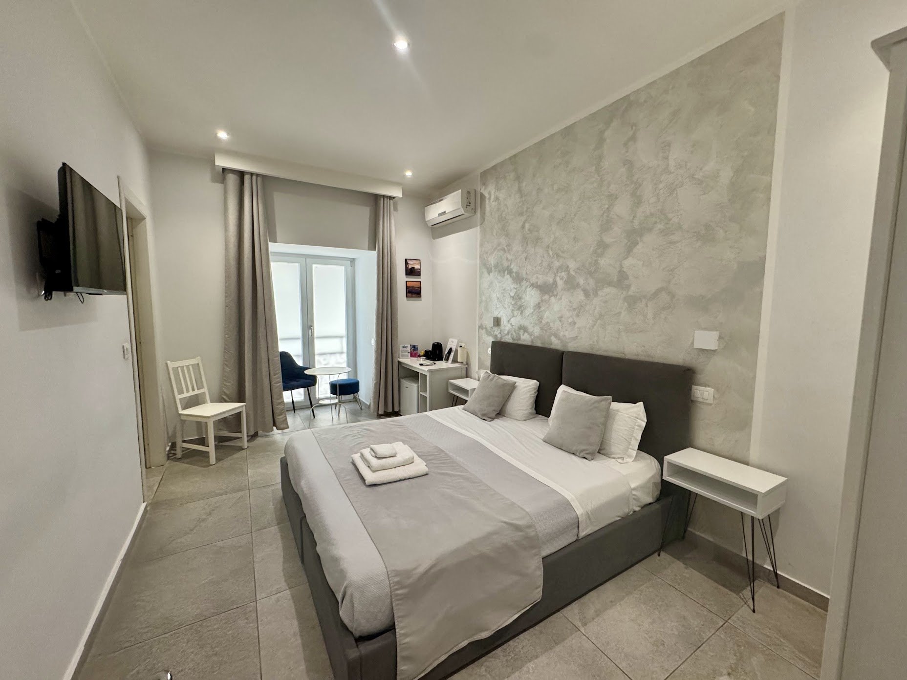 Hotel room with a bed, pillows, towels, a small white table, chairs, a TV mounted on the wall, window with curtains, air conditioning, and framed pictures.