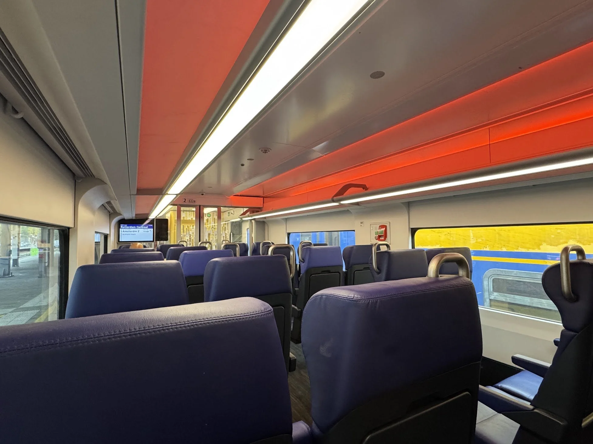 Interior of a train with purple seats, large windows, and a digital display showing destinations. The ceiling has red and white lighting.
