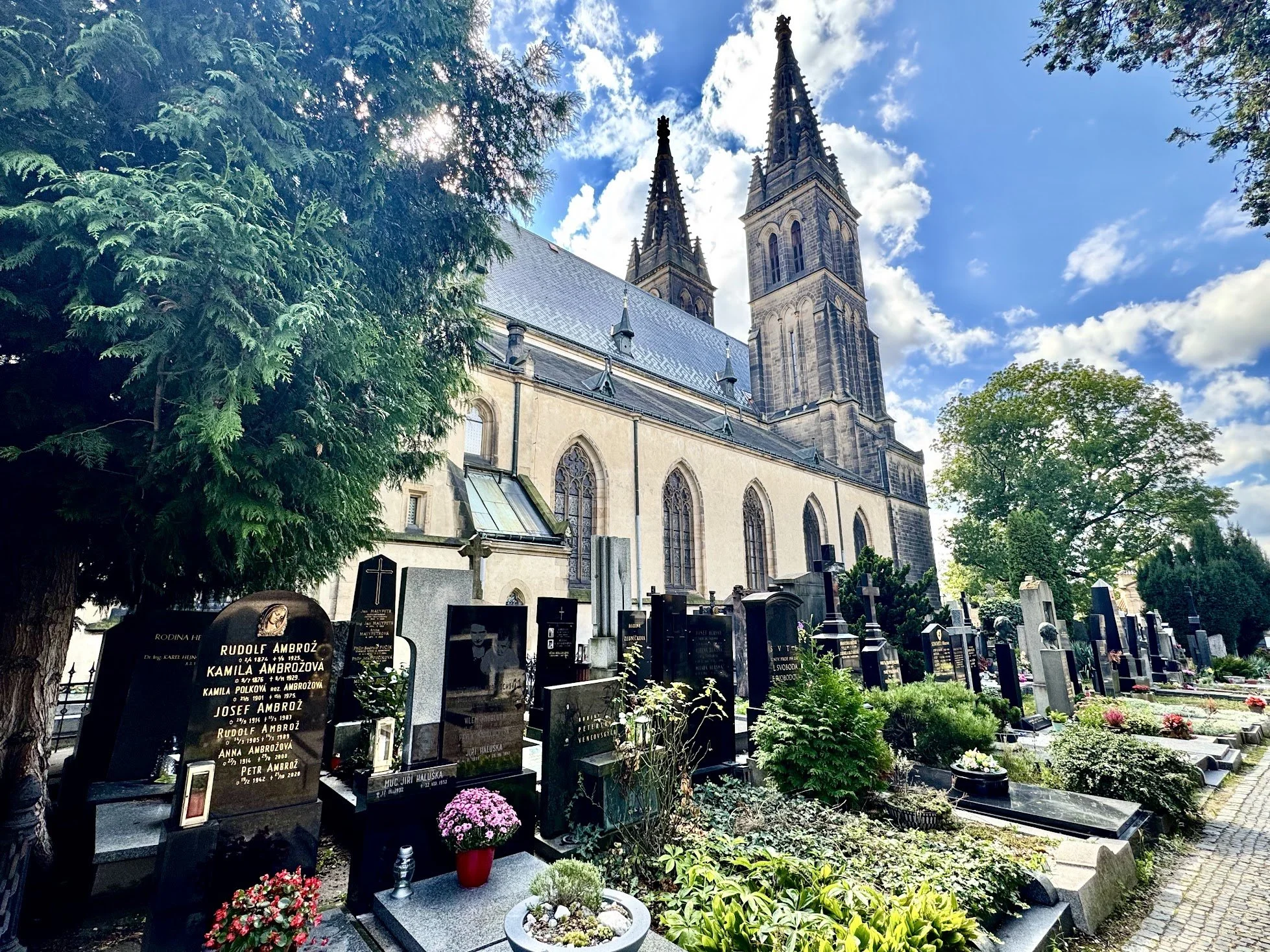 A large Gothic church with twin spires, surrounded by a graveyard with headstones and flowers, under a partly cloudy sky with sunlight filtering through the trees.