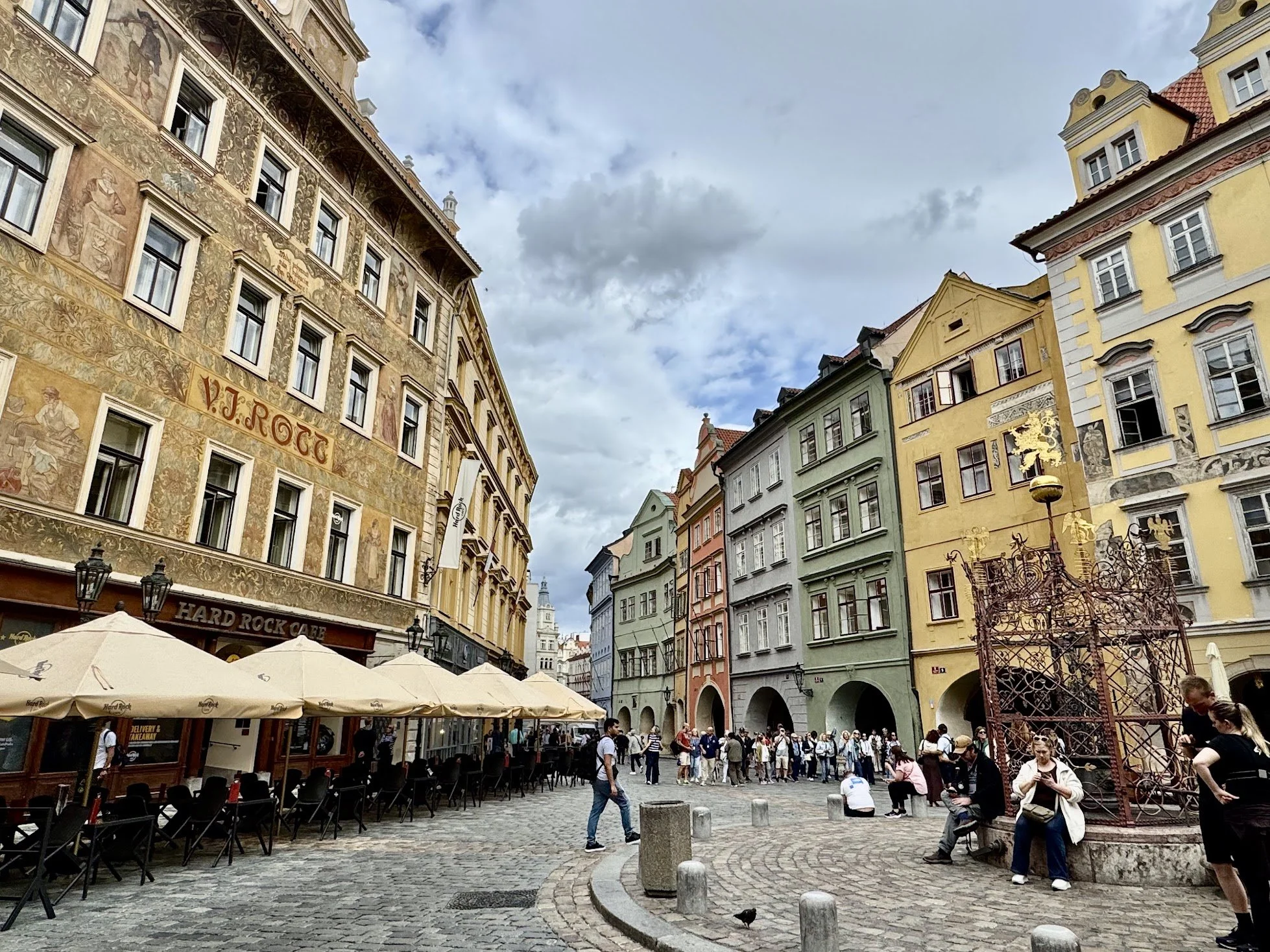 A bustling European square filled with people walking, sitting around outdoor cafes with umbrellas, and colorful historic buildings with ornate facades under a cloudy sky.