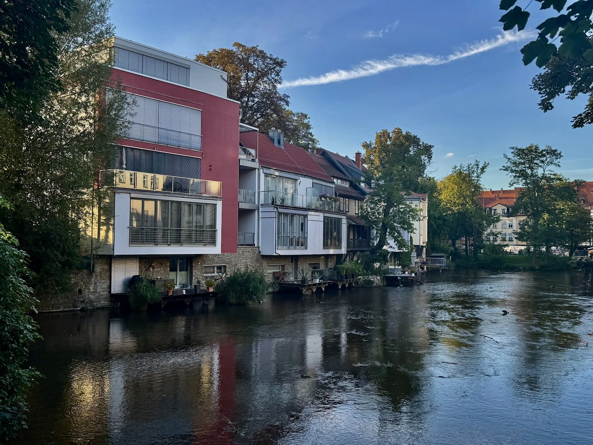 Residential buildings with modern architecture along a river, surrounded by trees and greenery, under a partly cloudy sky.