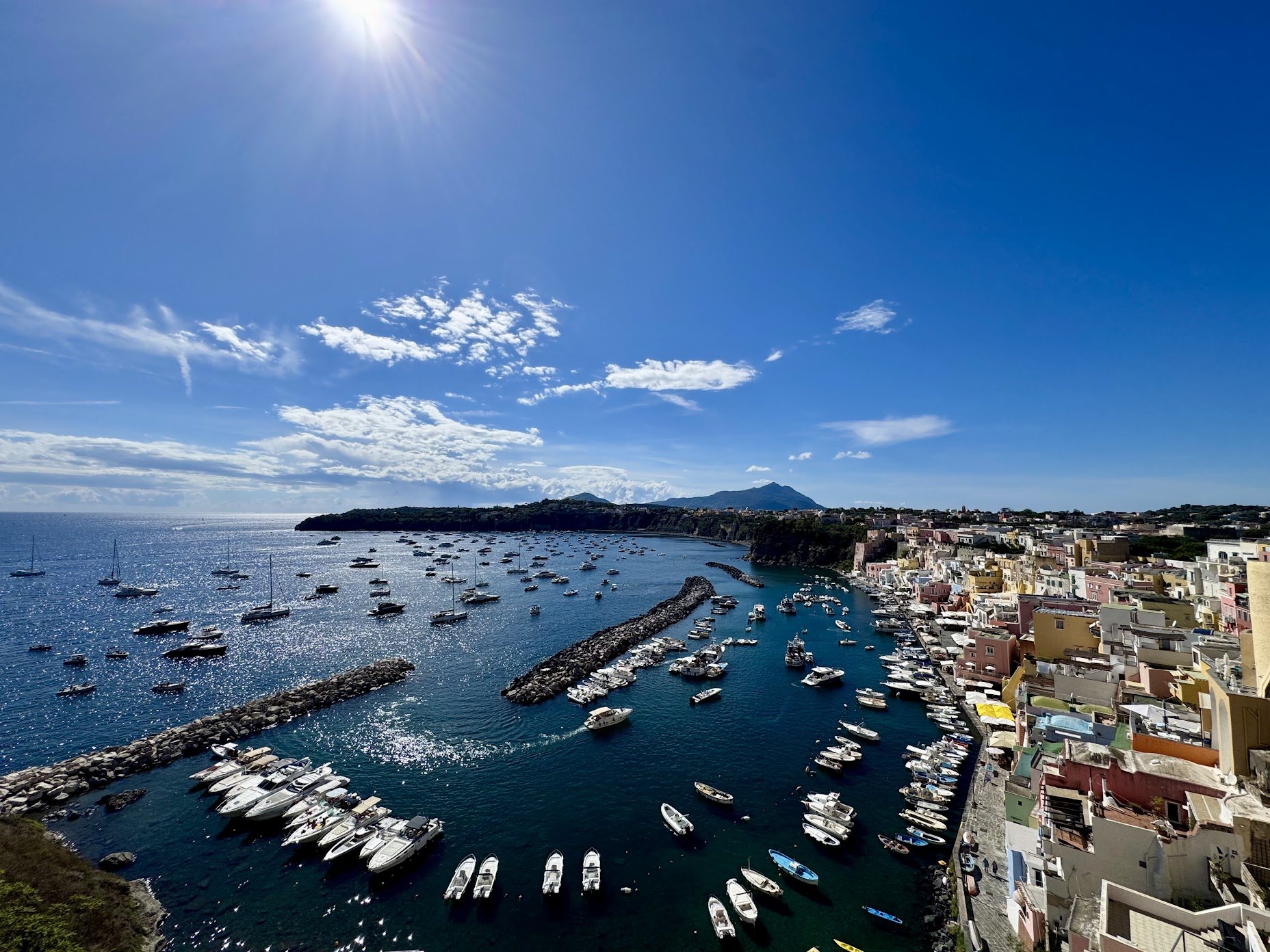 A coastal harbor with numerous boats and yachts docked, colorful buildings along the shoreline, mountains in the distance, and a bright blue sky with some clouds and the sun shining.