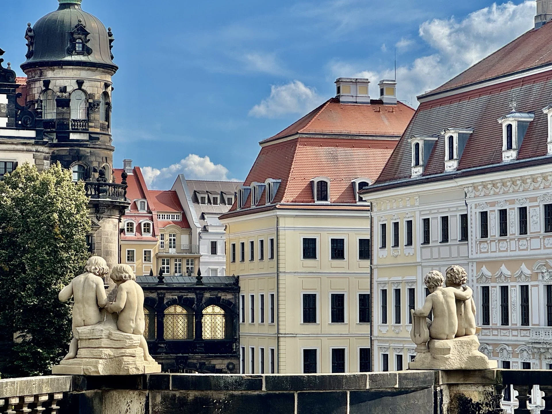 Statues of children on a stone balustrade in front of historic European buildings with mansard roofs and a cloudy blue sky.