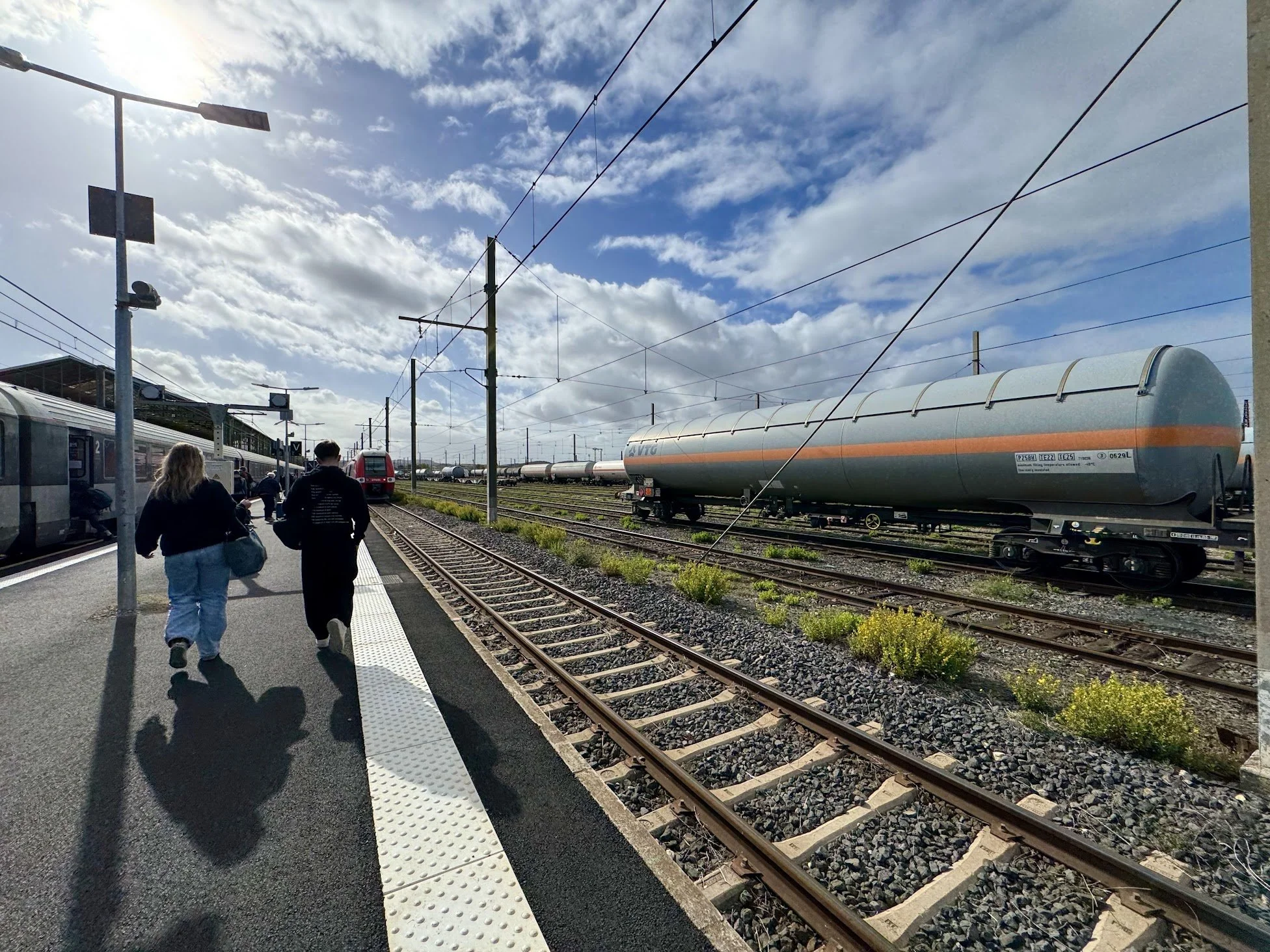 People walking on a train station platform with trains and train tracks, under a partly cloudy sky.