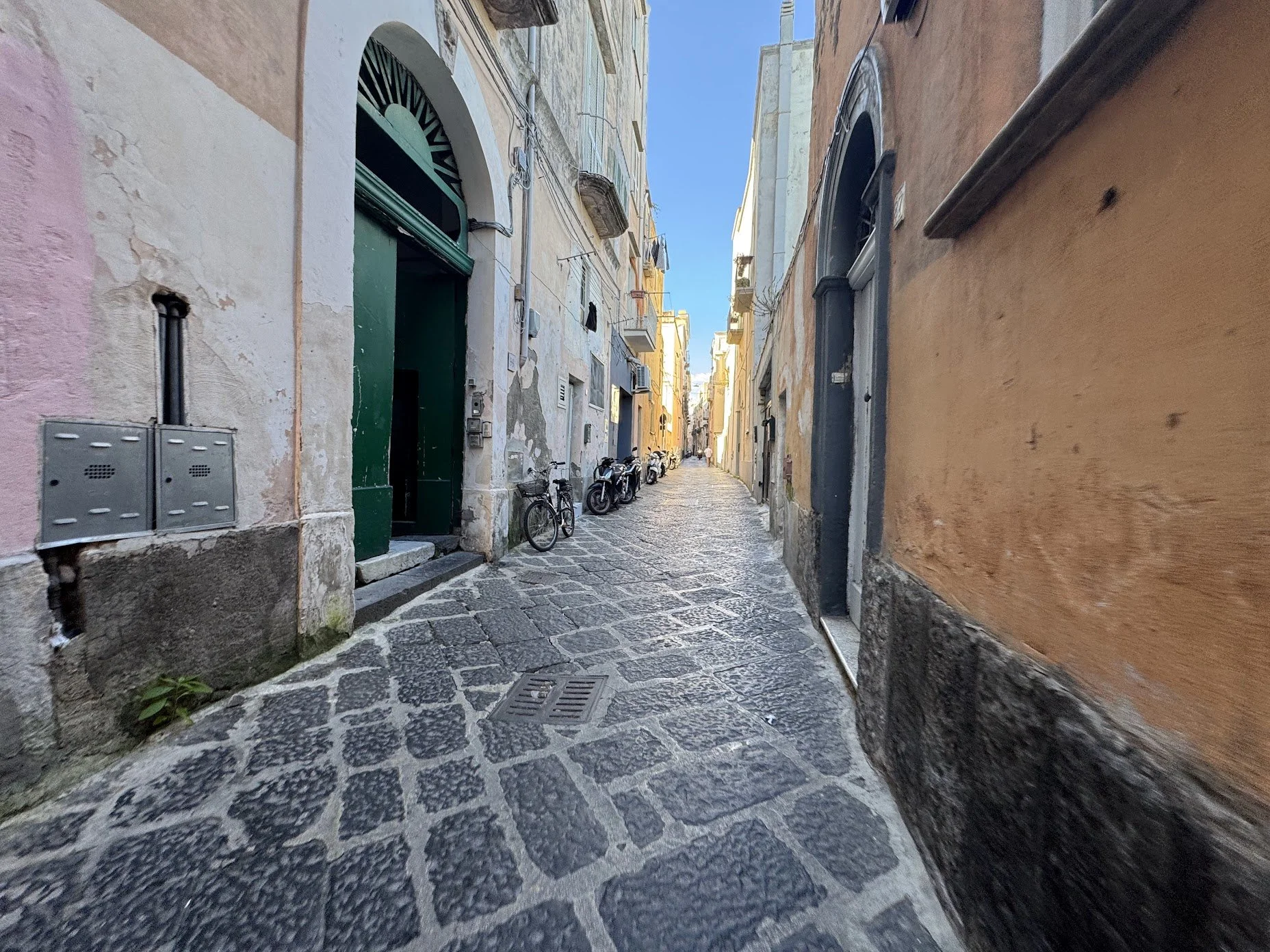 A narrow cobblestone alleyway in a European city, flanked by old, colorful buildings with various doors and windows, bicycles parked along the side, under a clear blue sky.