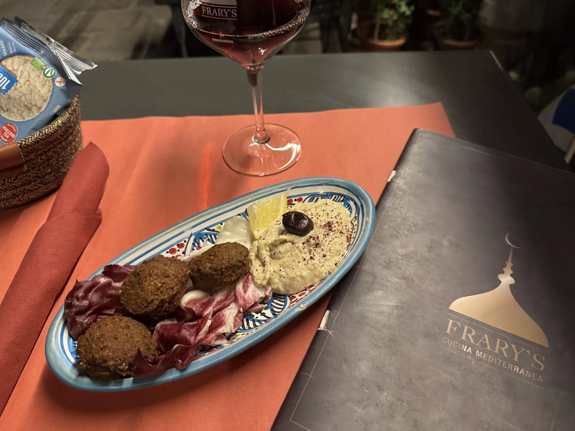 A table setting with a glass of red wine and a colorful dish of Mediterranean cuisine including falafel, hummus, lemon wedge, olives, and roasted vegetables on a blue patterned plate.
