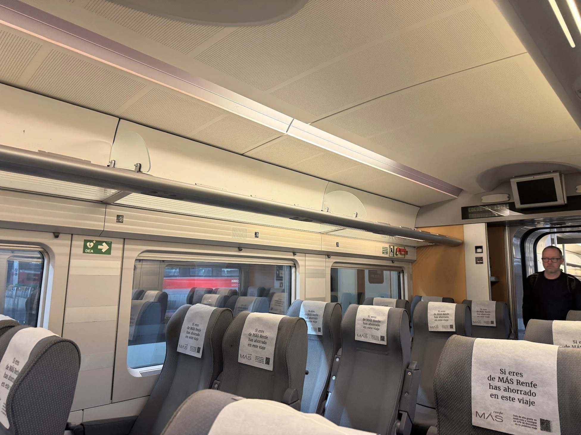 Interior of a train with empty gray seats, some with paper signs reading in Spanish, indicating passengers have saved the seats. Overhead compartments and a small monitor are visible at the front.