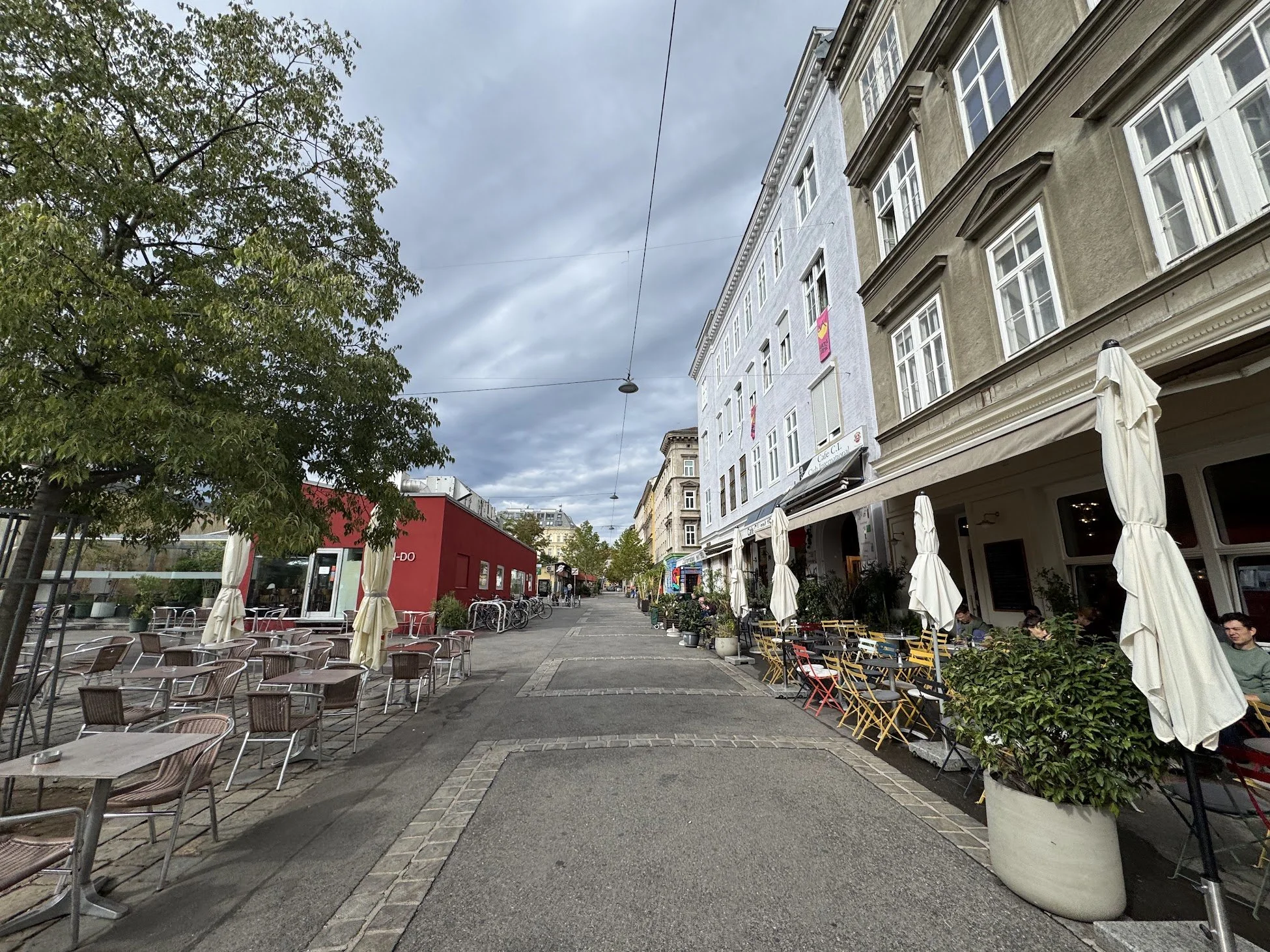 Outdoor street cafe with tables and chairs, surrounded by large potted plants, with buildings and cloudy sky overhead.