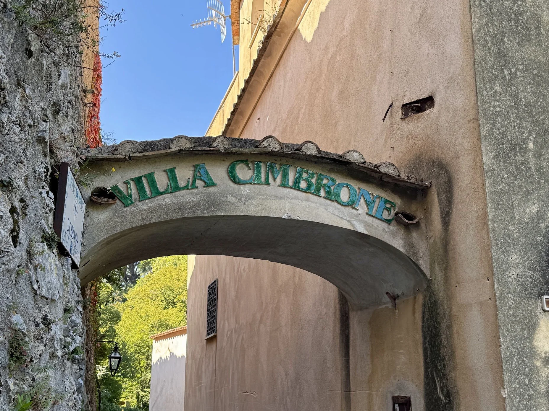 Archway with a sign reading 'Villa Cimbrome' in green letters, stone wall on the left, building with a pinkish hue behind, and trees with green leaves in the background.