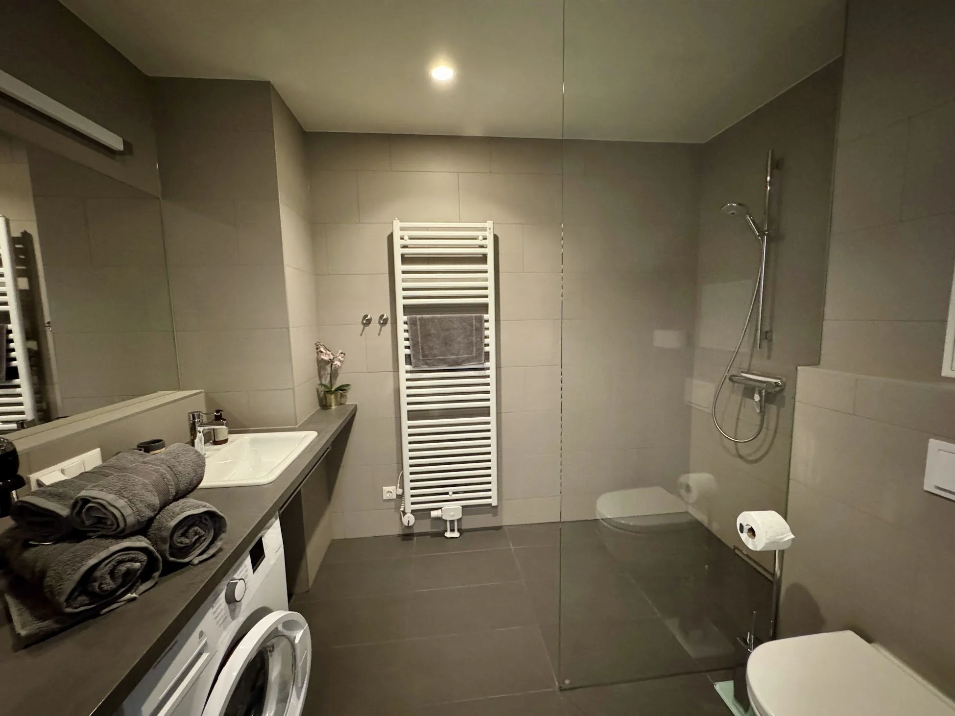 Modern bathroom with gray tiled walls and floor, a white towel radiator, a shower area with a handheld showerhead, a white toilet, a washbasin with hand towels, and a row of black towels on a countertop.