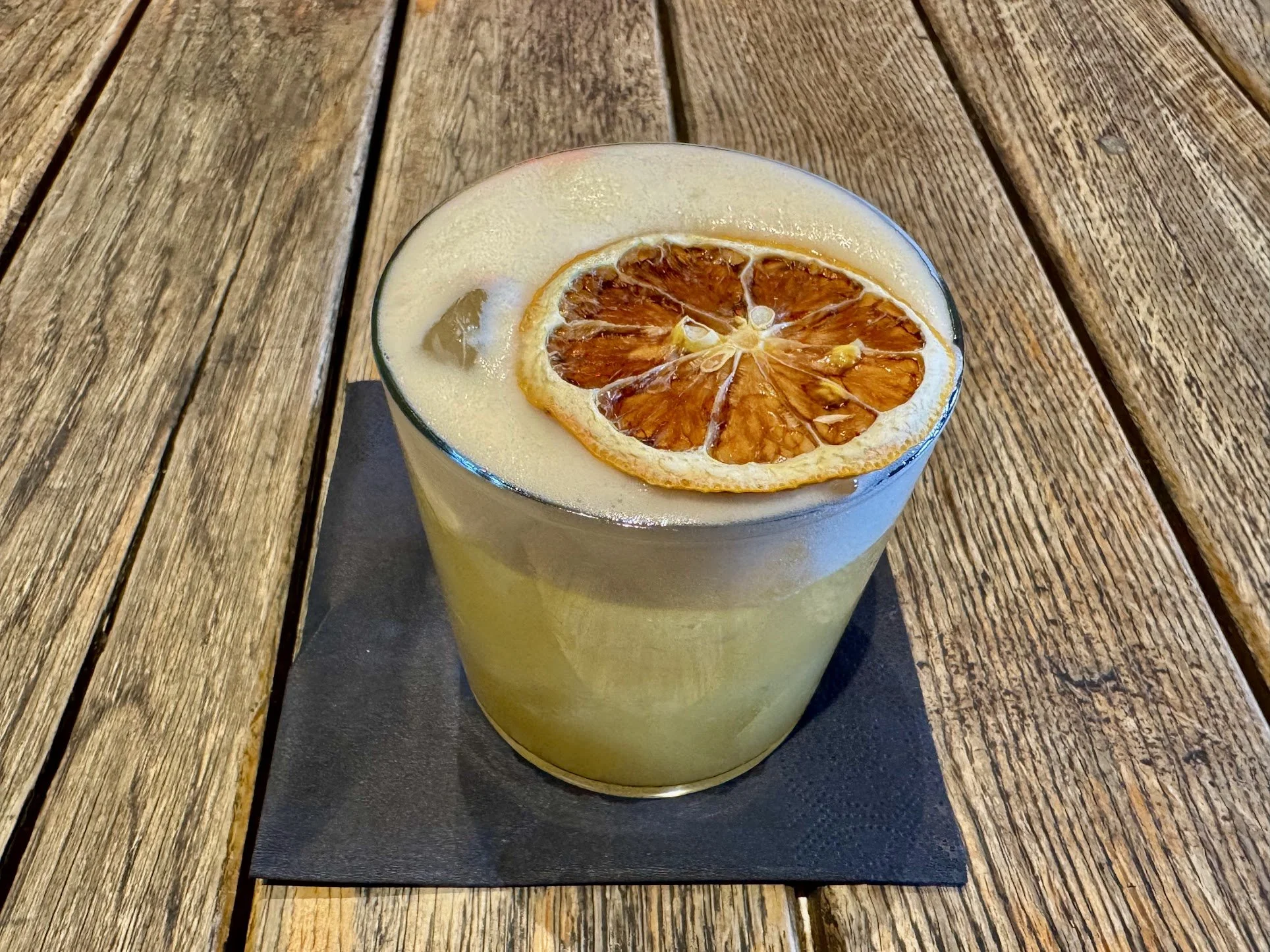 A glass of lemonade with a slice of dried orange on top, placed on a black napkin on a wooden surface.