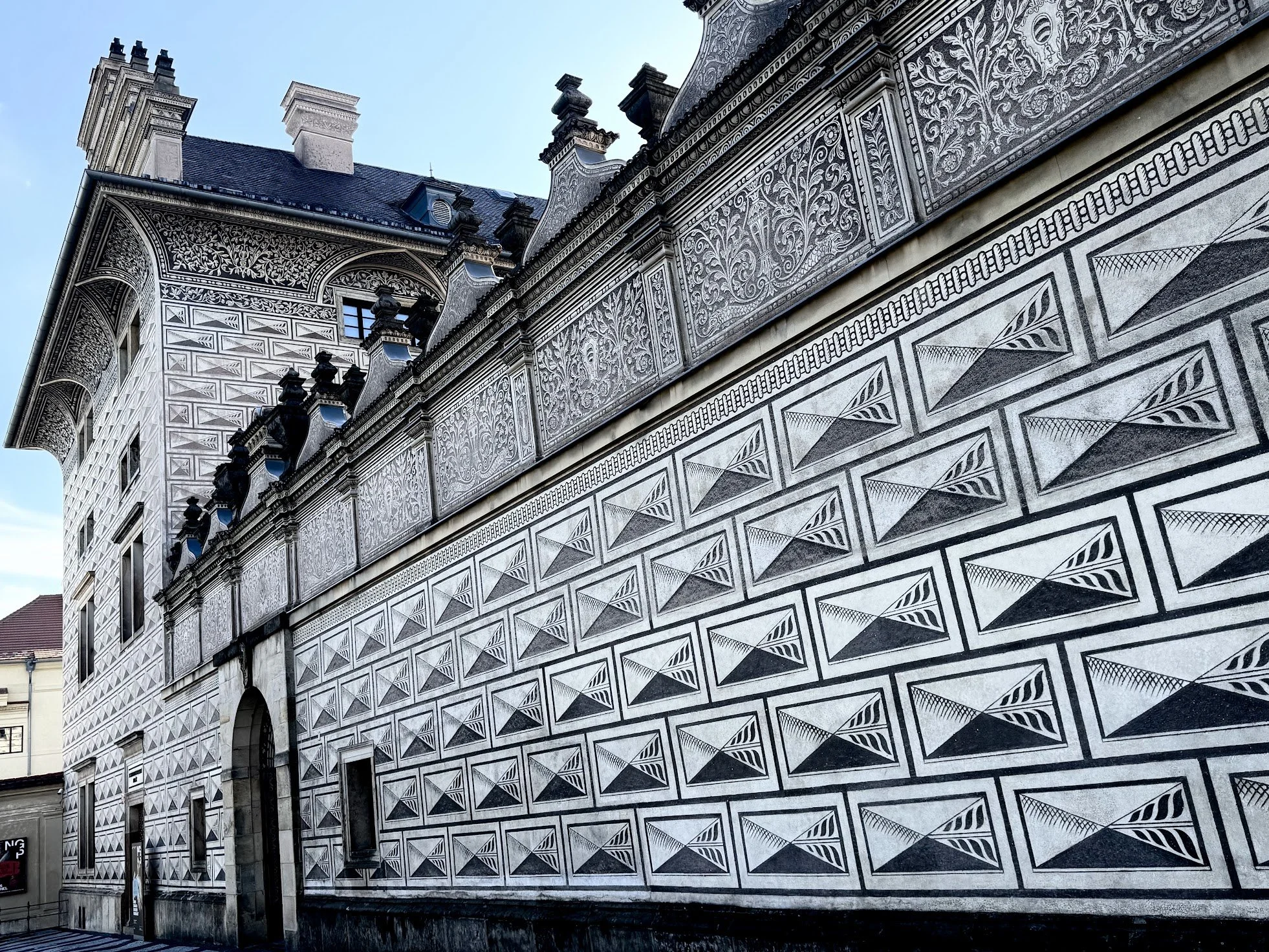 Close-up view of a historic building facade with intricate black and white geometric and floral patterns, including arched windows and decorative chimneys against a clear blue sky.