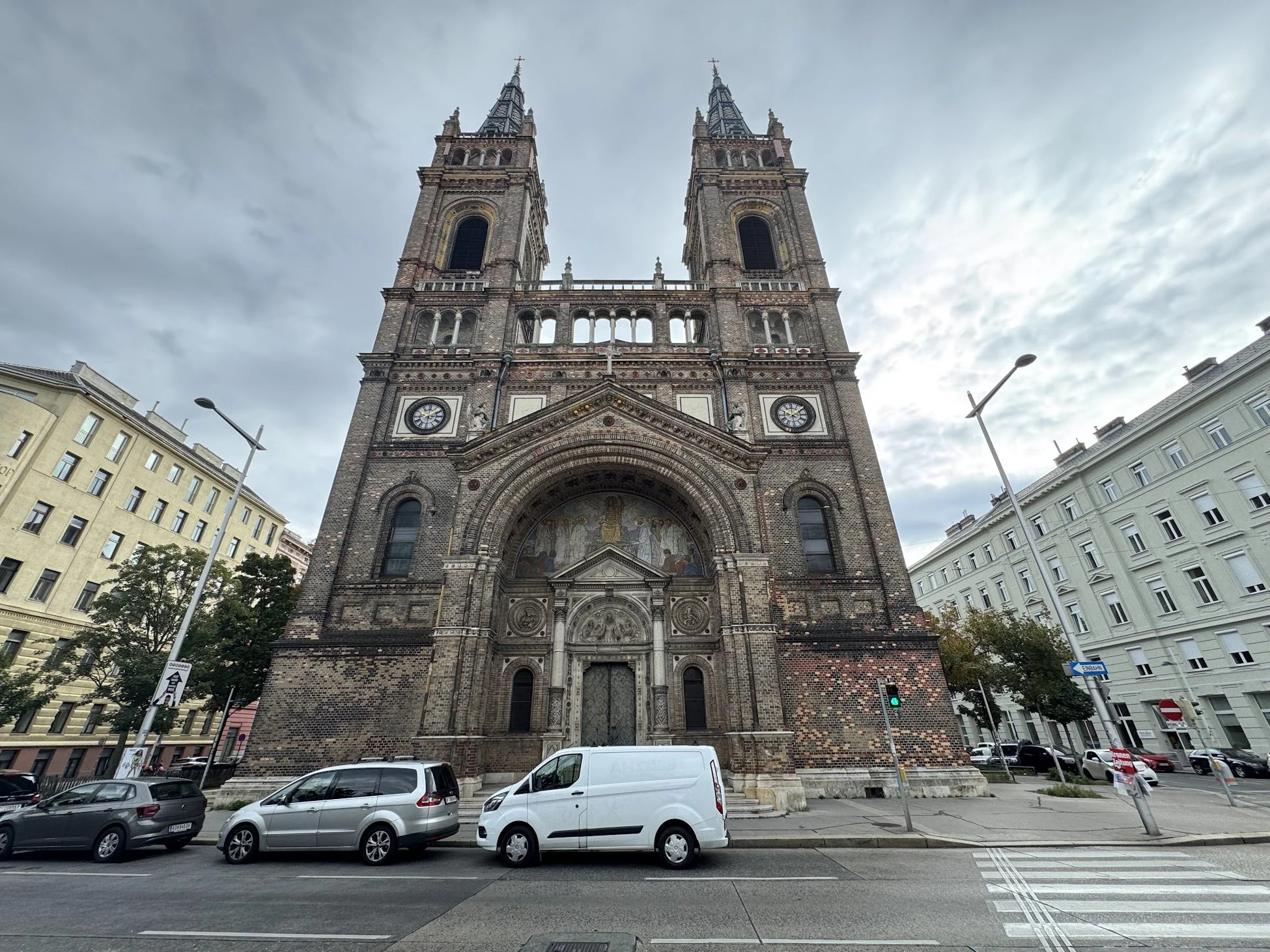 Front view of a historic church with twin towers, decorative brickwork, and a large arched entrance, located in an urban setting with parked cars, surrounding buildings, and street signs.