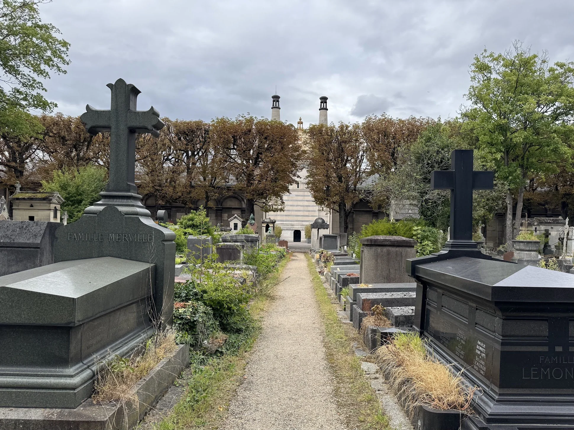 A cemetery with tombstones and crosses, some engraved with family names, surrounded by trees and greenery, with a cloudy sky overhead.