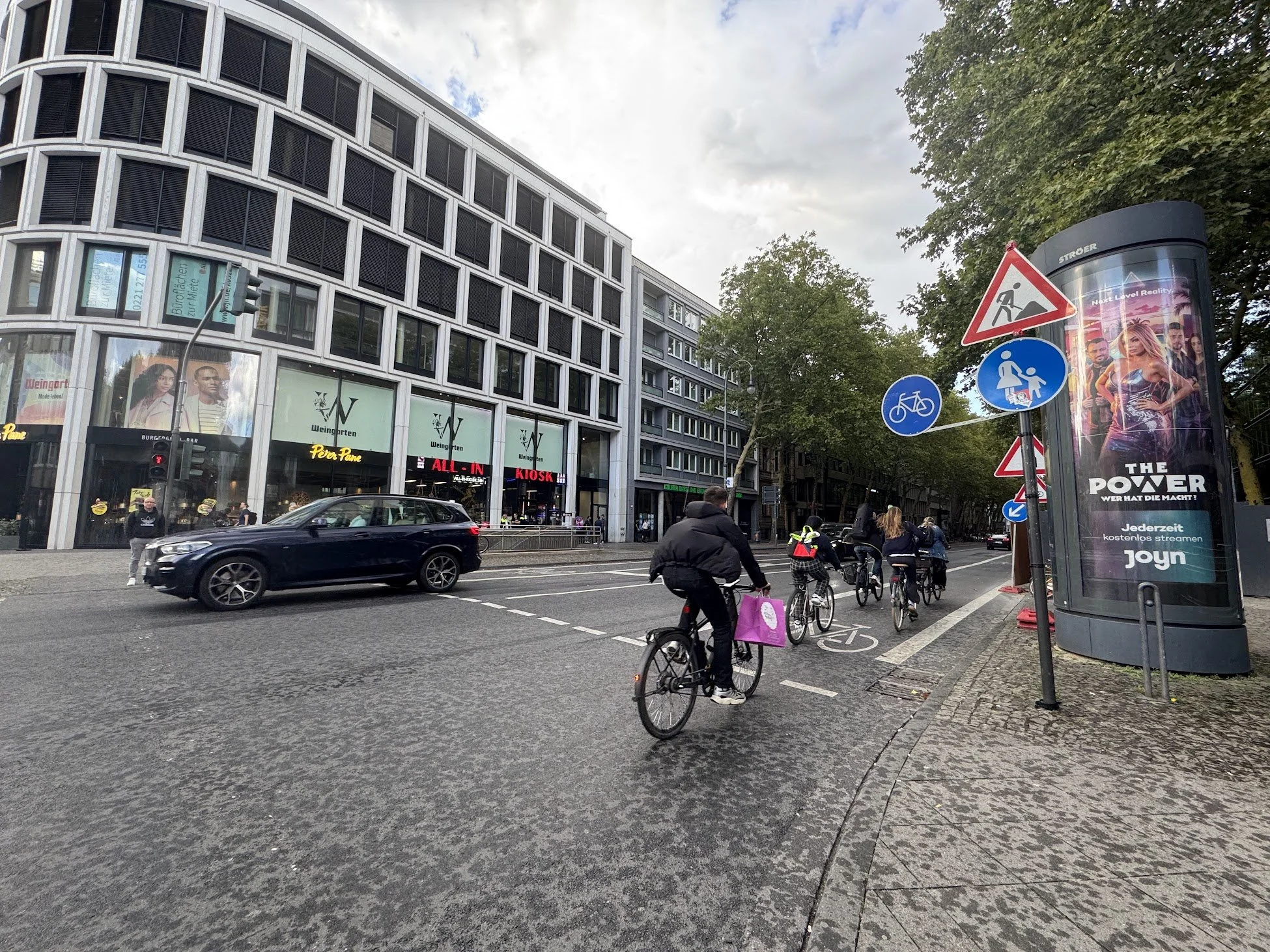 Urban street scene with a modern building, people riding bicycles and walking, cars on the road, and street signs indicating a bicycle lane and pedestrian crossing.