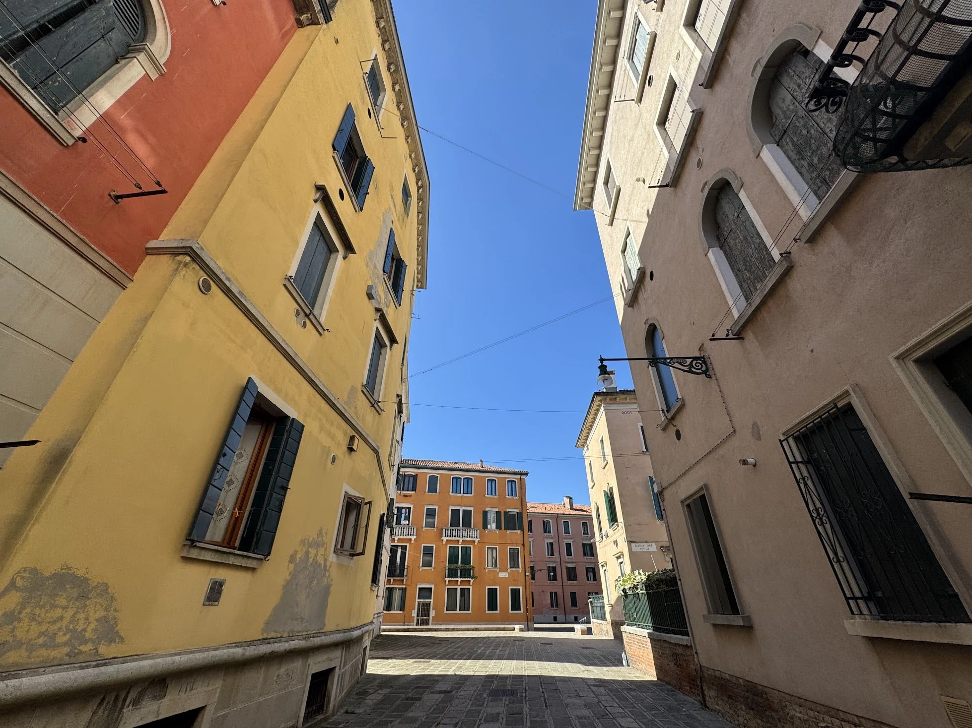 A narrow urban courtyard surrounded by tall colorful buildings with small balconies and green shutters, under a clear blue sky.