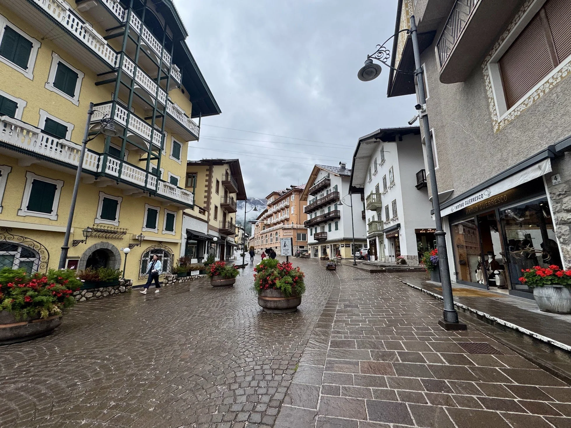A European-style pedestrian street with colorful buildings, flower pots, and people walking on wet cobblestone pavement, under a cloudy sky.