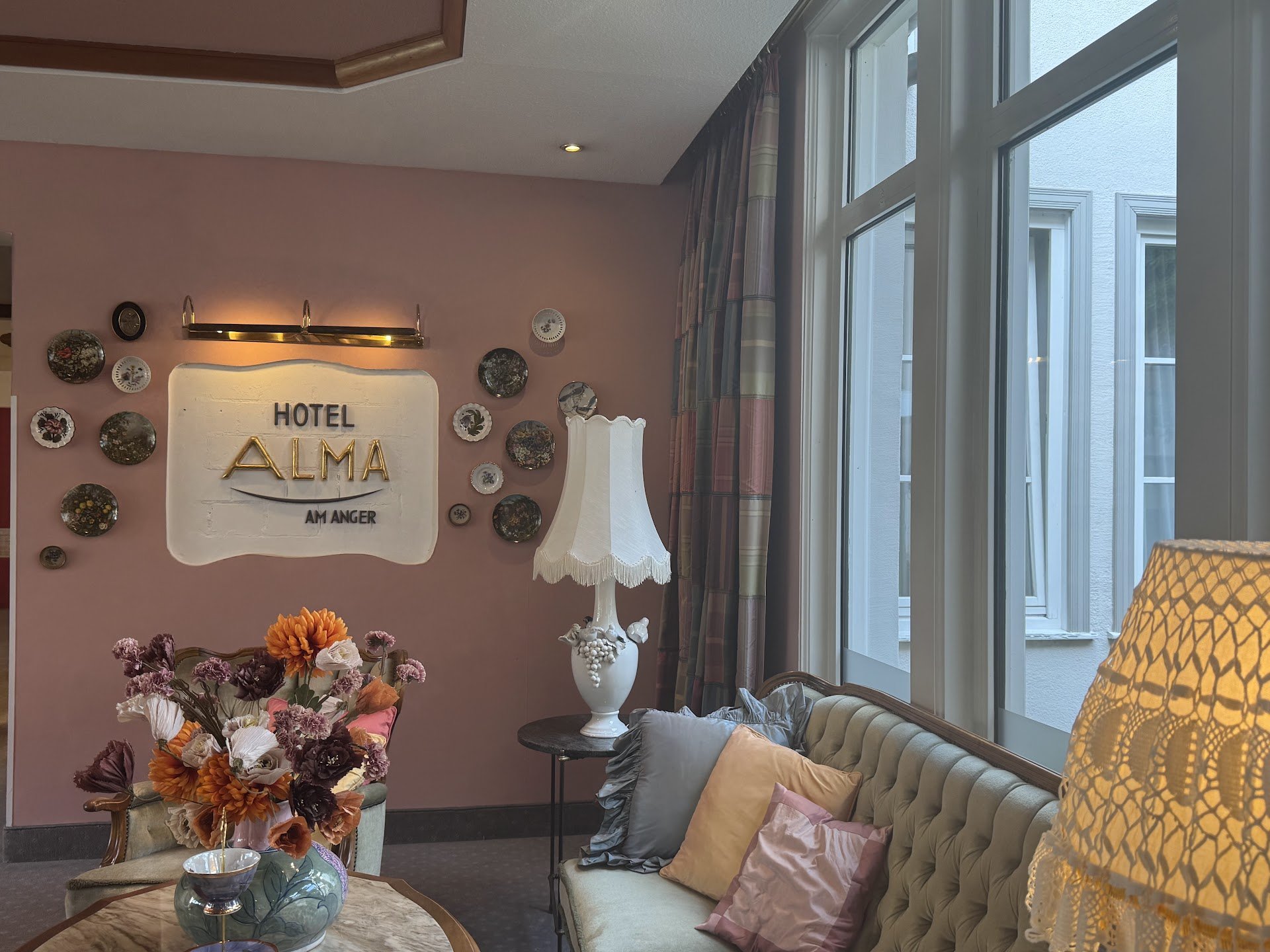 Interior of a hotel room with pink wall, decorated with plates and a sign reading 'Hotel Alma, Amnger.' There are large windows with plaid curtains, a cream-colored tufted sofa with pillows, an ornate white lamp, and a vase of colorful flowers on a m