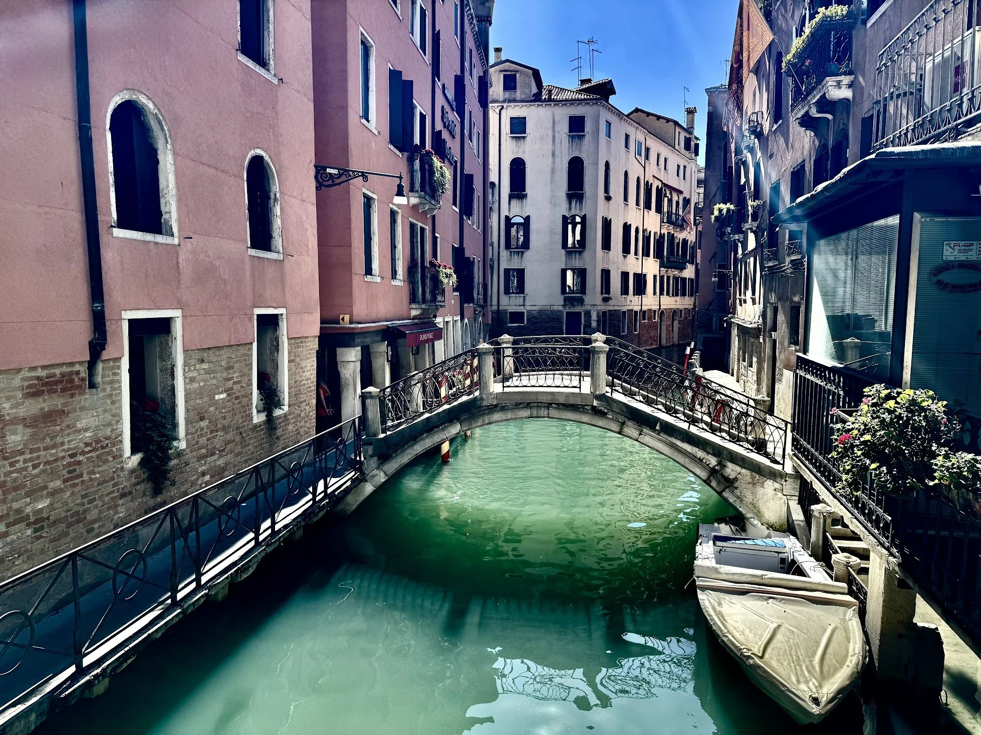 A narrow canal with green water flowing under a small bridge, surrounded by tall buildings with colorful facades and balconies, in Venice, Italy.