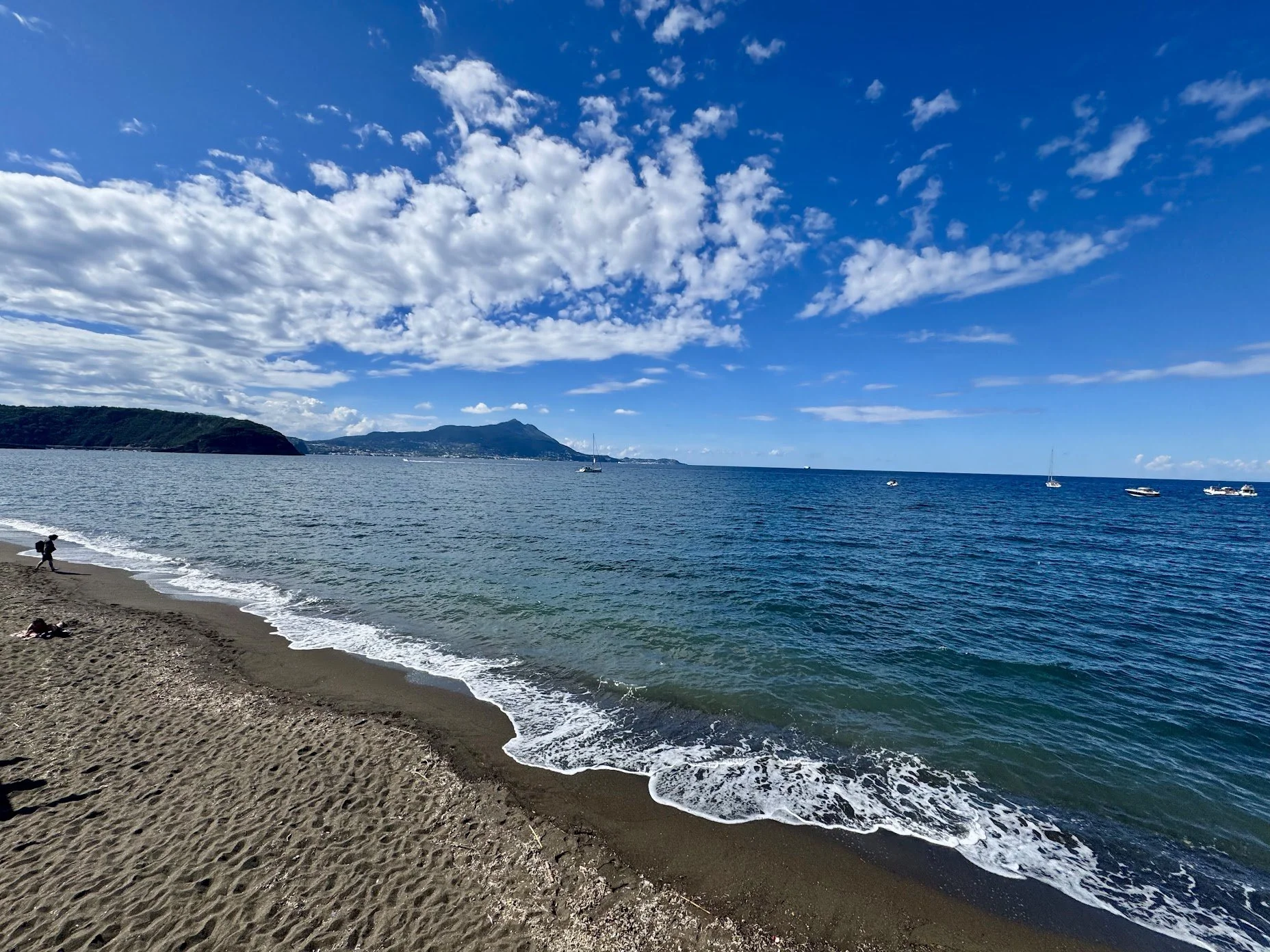 A beach with sand, small waves, and a few boats floating in calm blue water, with a hilly landmass in the background under a partly cloudy sky.